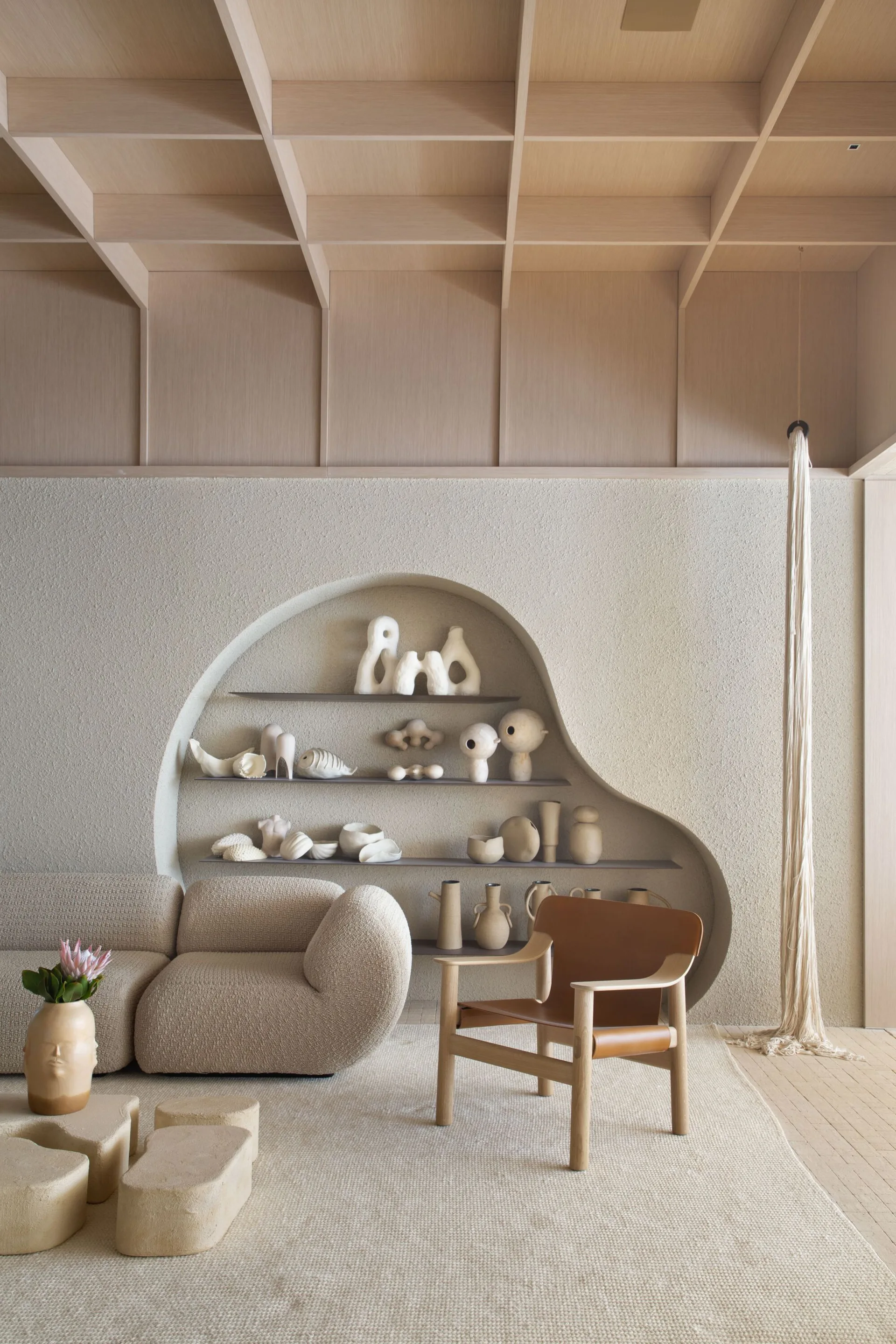 A neutral living room featuring a curved plaster wall alcove with shelves, a textured boucle sofa, and a coffered wood ceiling.