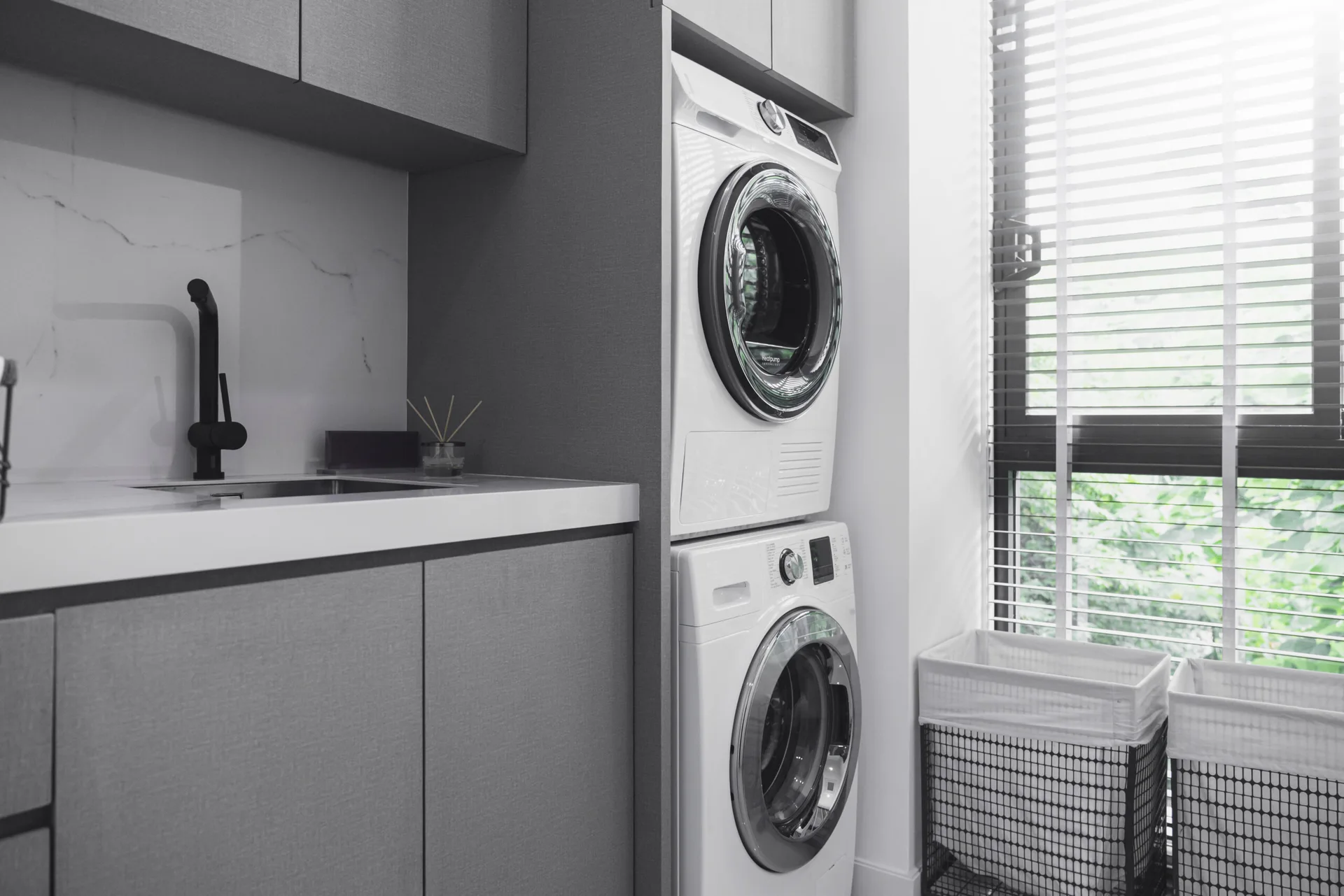 A modern laundry room featuring stacked white appliances, matte grey cabinets, an undermount sink with a black faucet, and a window with blinds.