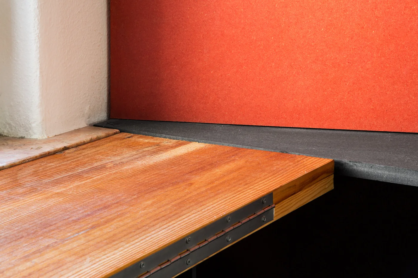 A close-up detail of a wooden panel with a metal piano hinge abutting a dark grey stone ledge beneath a bright orange wall.