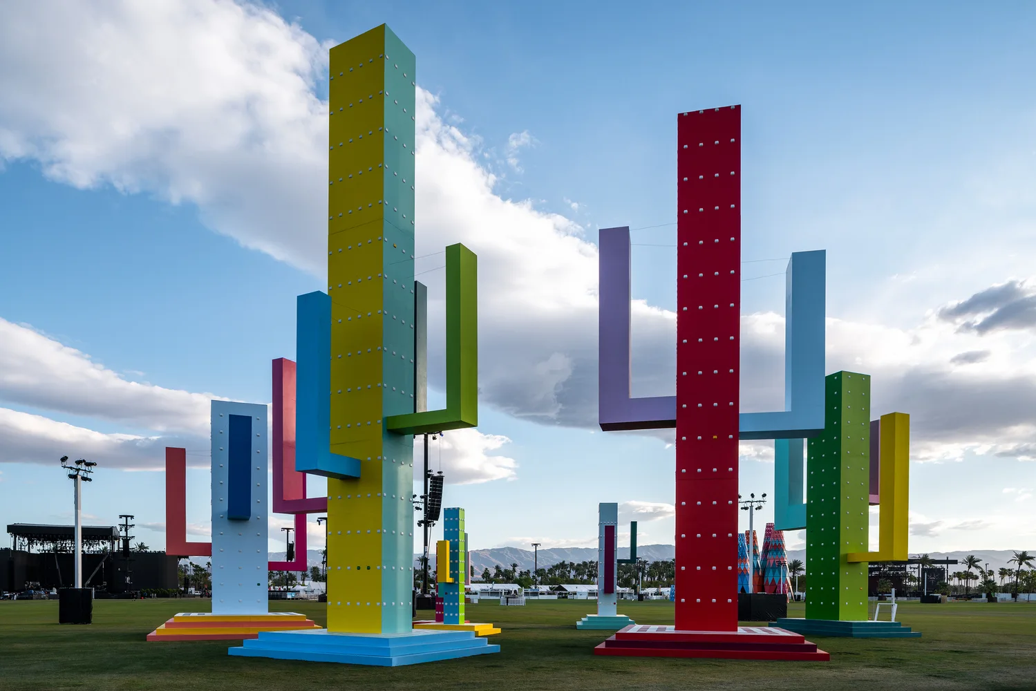 Tall, colorful cactus-shaped geometric sculptures stand on an open grassy field under a cloudy sky.