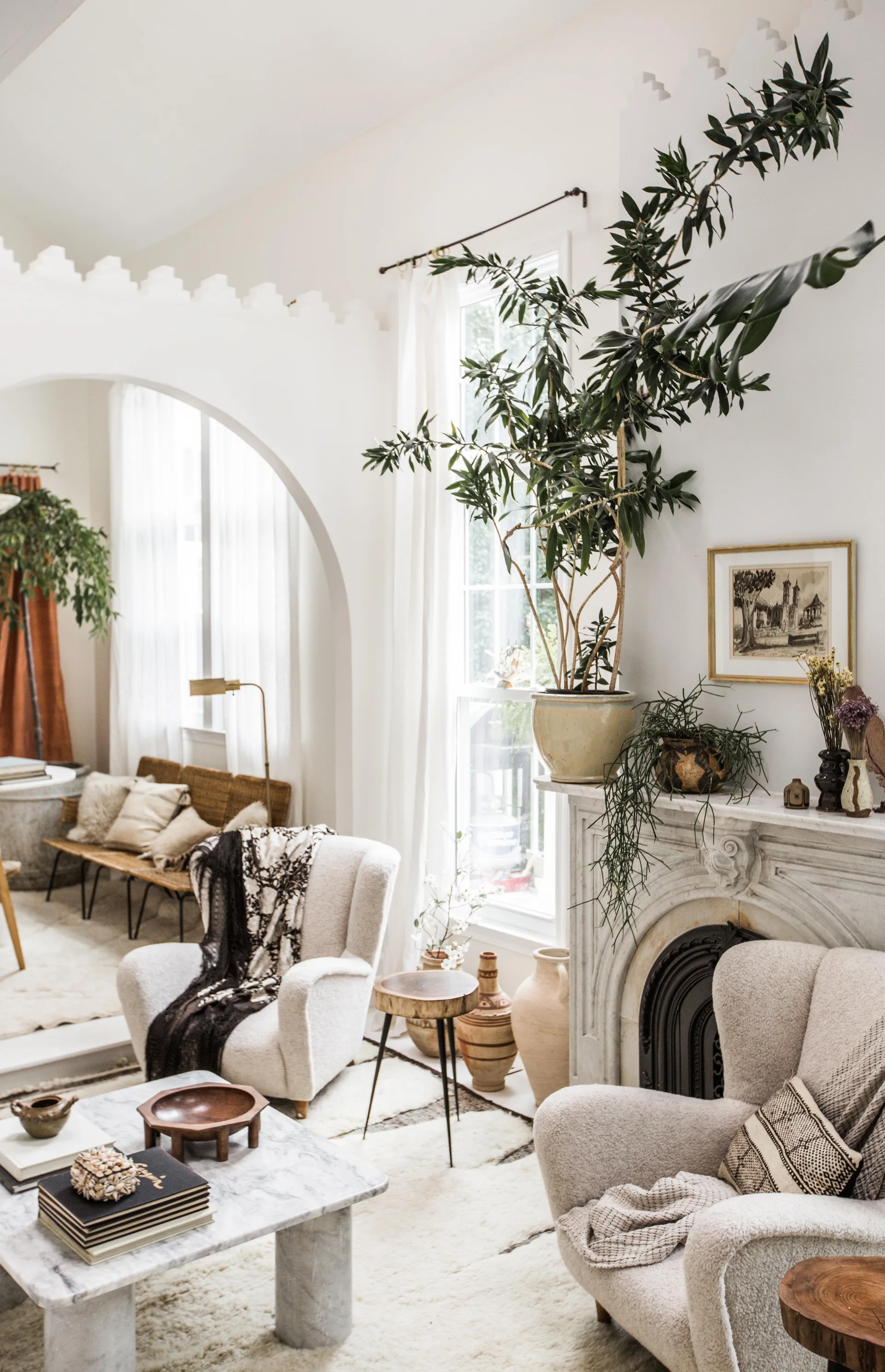 A bright living room features two plush cream armchairs, a marble fireplace with potted plants, and an architectural archway leading to another room.