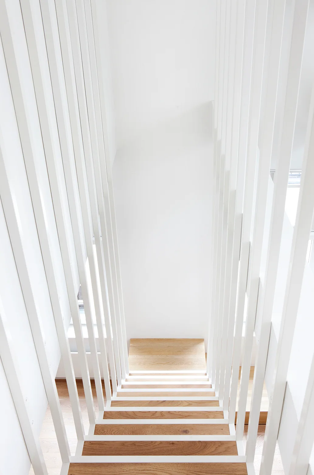 Looking down a minimalist stairwell featuring light wood treads framed by tall vertical white metal balusters.