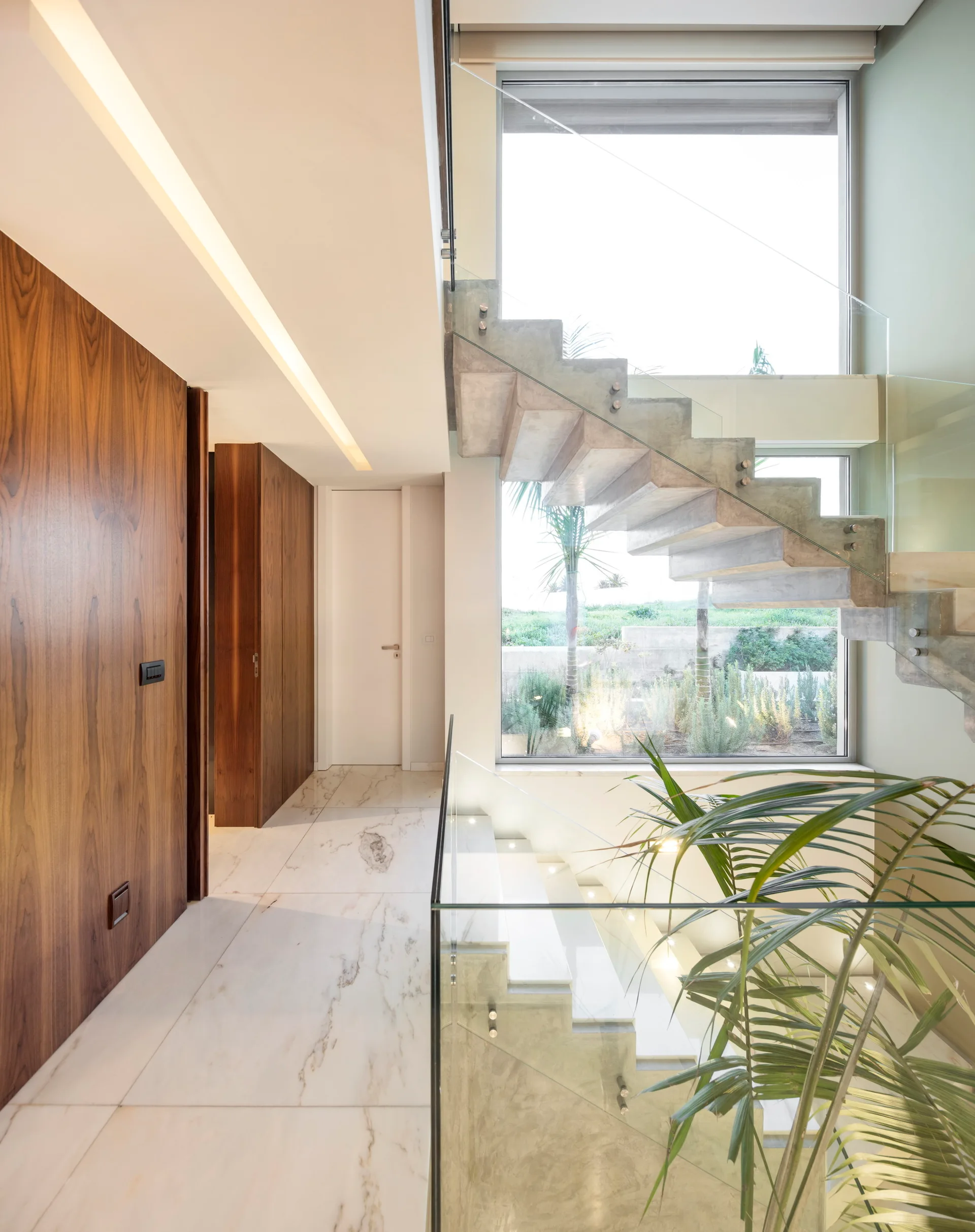 A modern staircase with floating concrete steps and glass railings sits next to rich wood wall paneling and a polished marble floor.