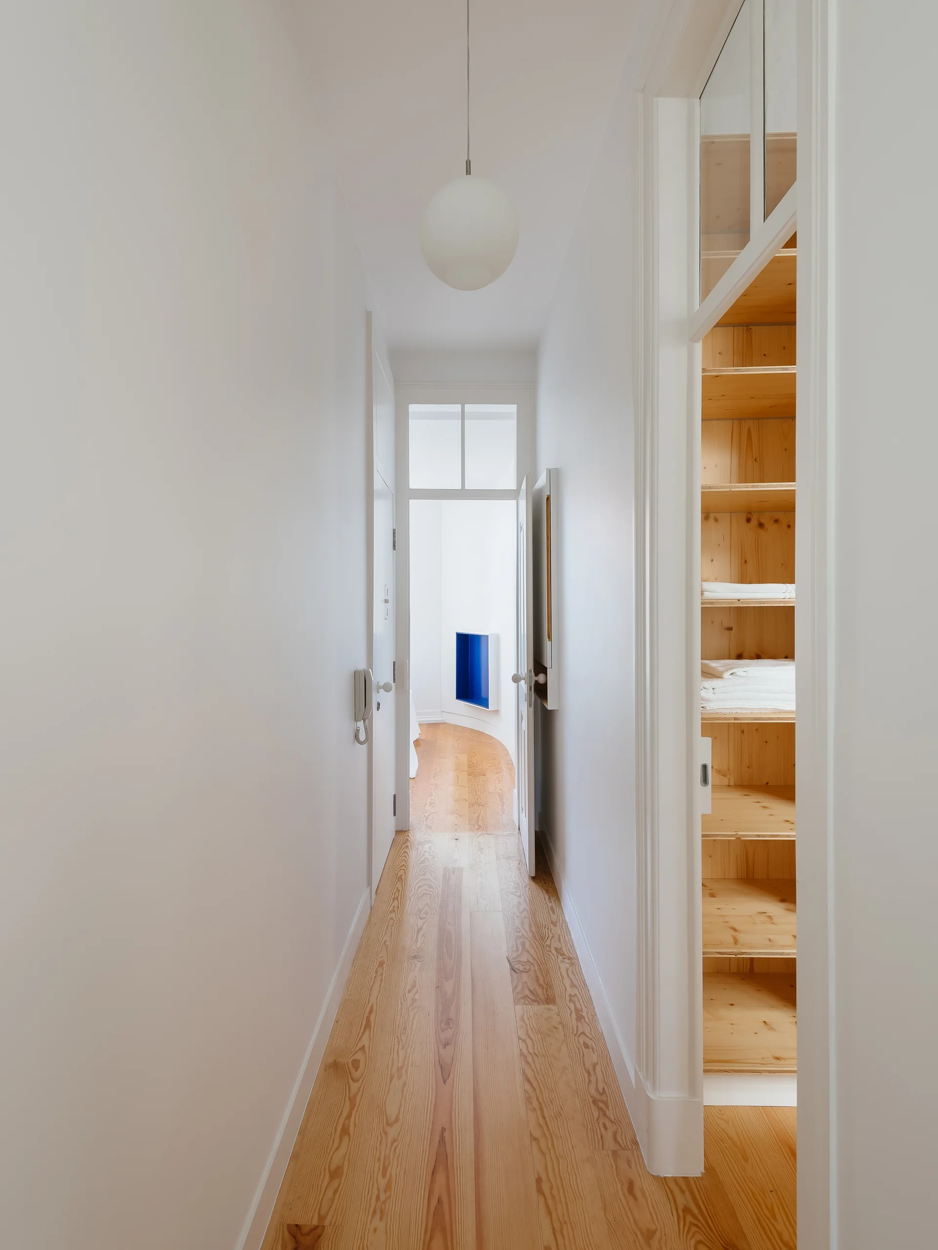 A narrow white hallway features light-toned wood floors, a hanging globe pendant light, and an open closet showing natural wood shelves filled with linens.
