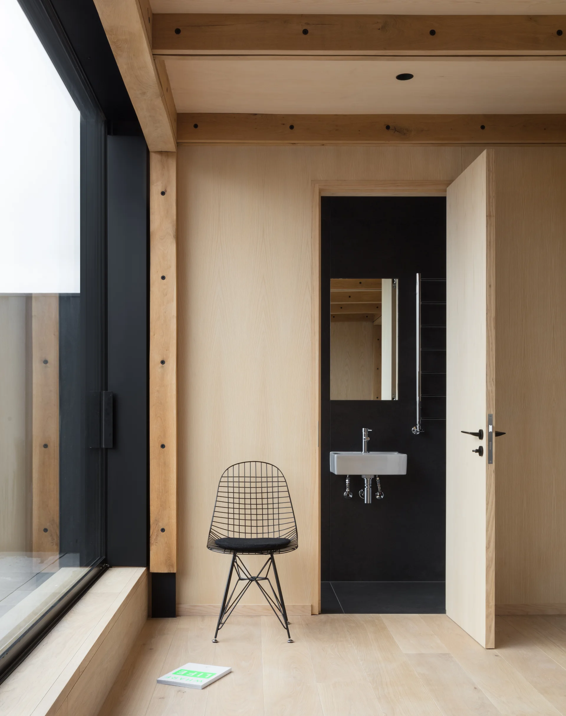 A minimalist room with light wood walls and a black wire chair features an open doorway to a matte black bathroom with a white sink.