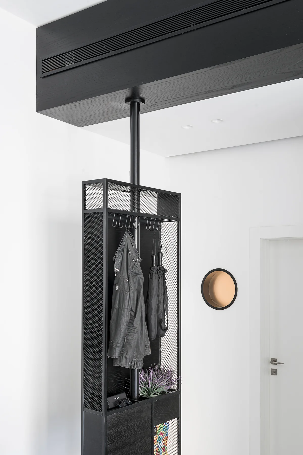 A tall black metal floor-to-ceiling coat rack with mesh panels stands in a white entryway next to a circular wall niche.