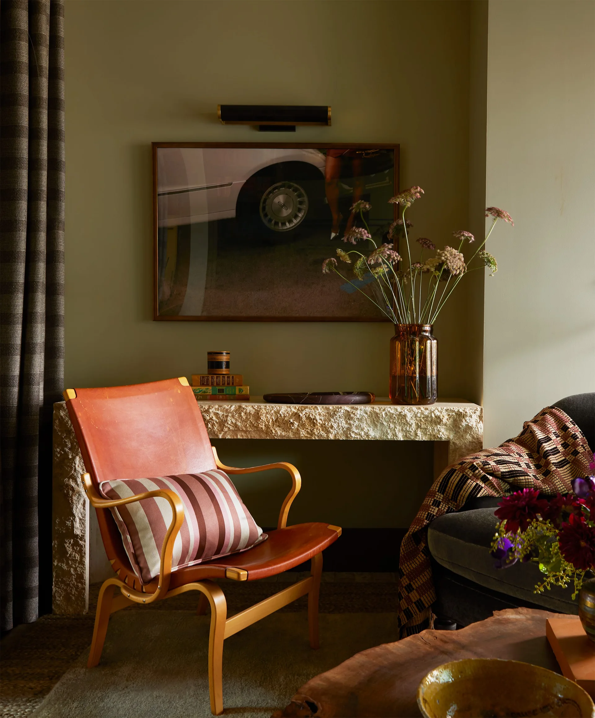 A leather lounge chair with a striped pillow sits in front of a rough stone console table against an olive green wall.