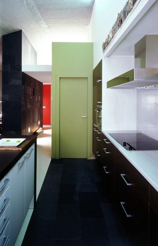 A modern galley kitchen features dark wood cabinets, a black tiled floor, and a bright green partition wall with a matching door.