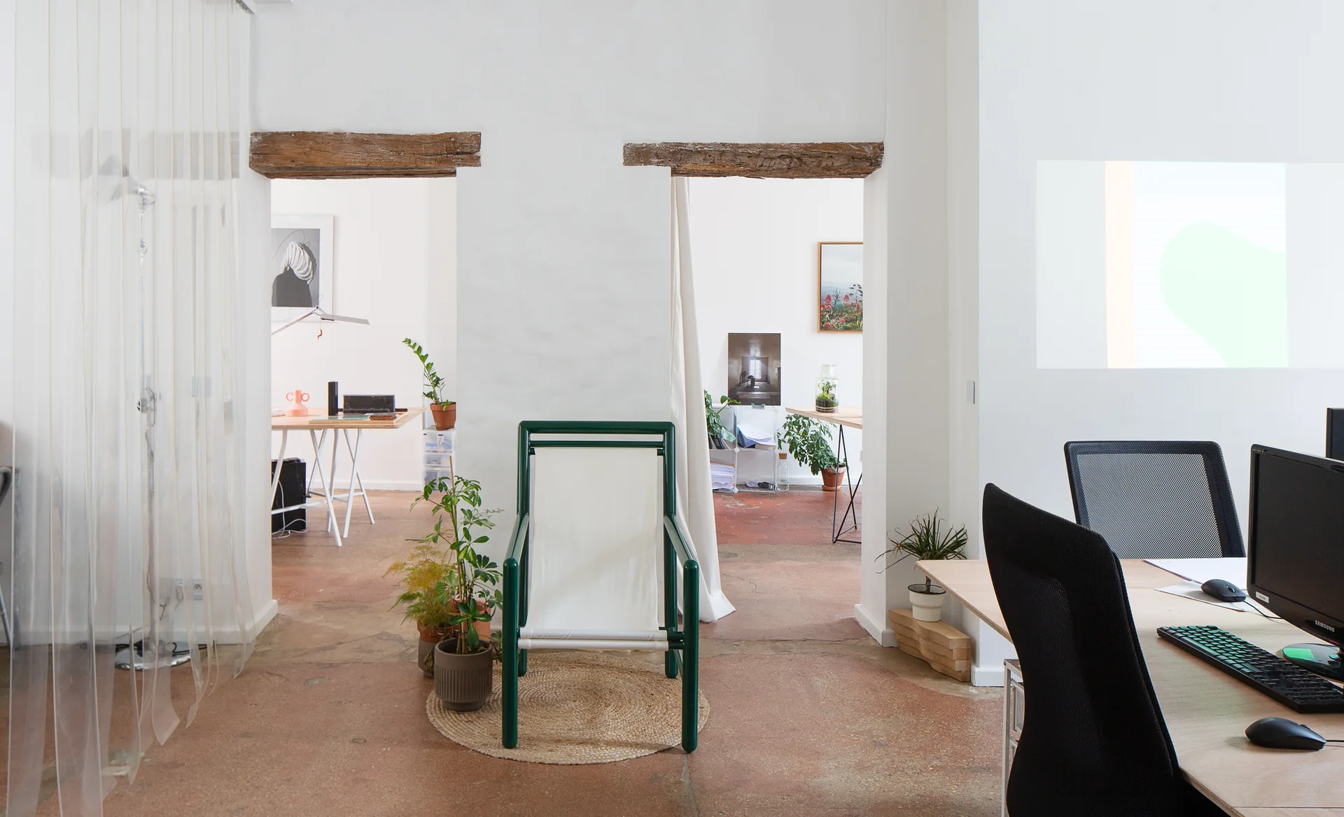 An open office space features white walls, terracotta floors, two doorways with rustic wood lintels, and a green armchair on a round jute rug.