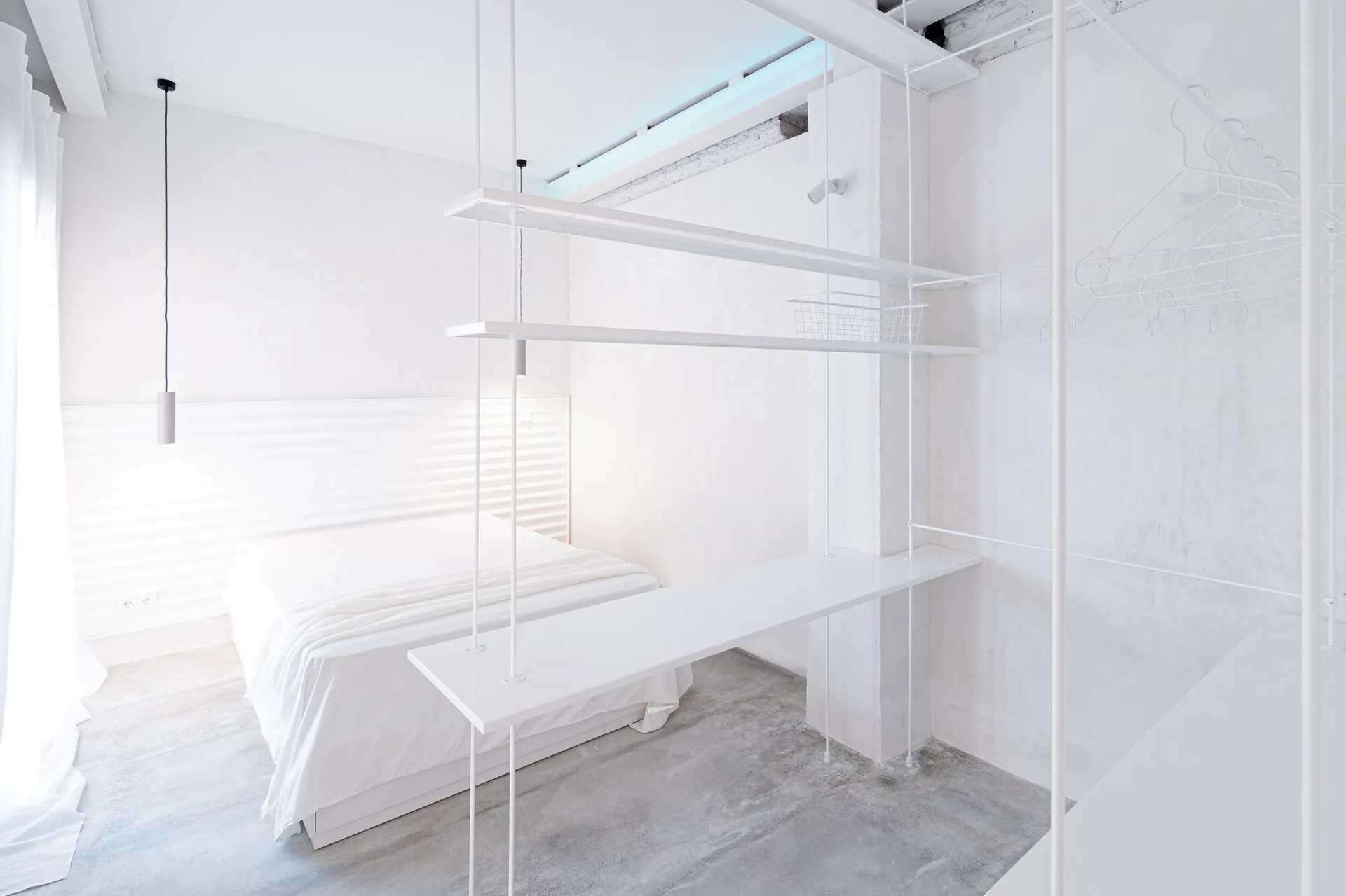 A minimalist white bedroom features a platform bed, a custom suspended white metal shelving system, and a smooth concrete floor.