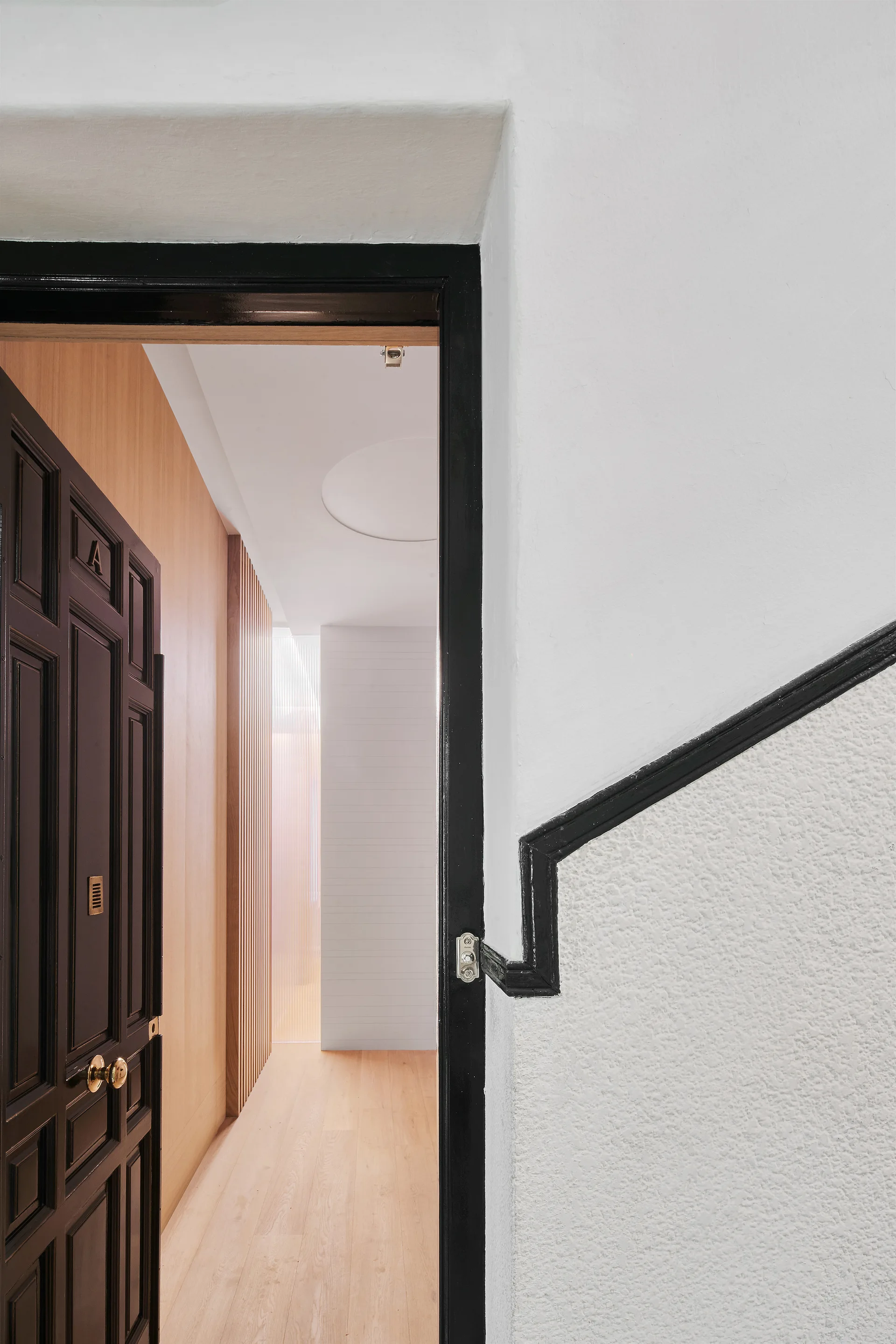 A view through an open black door into a bright hallway with light wood floors, white walls, and vertical wood slat paneling.