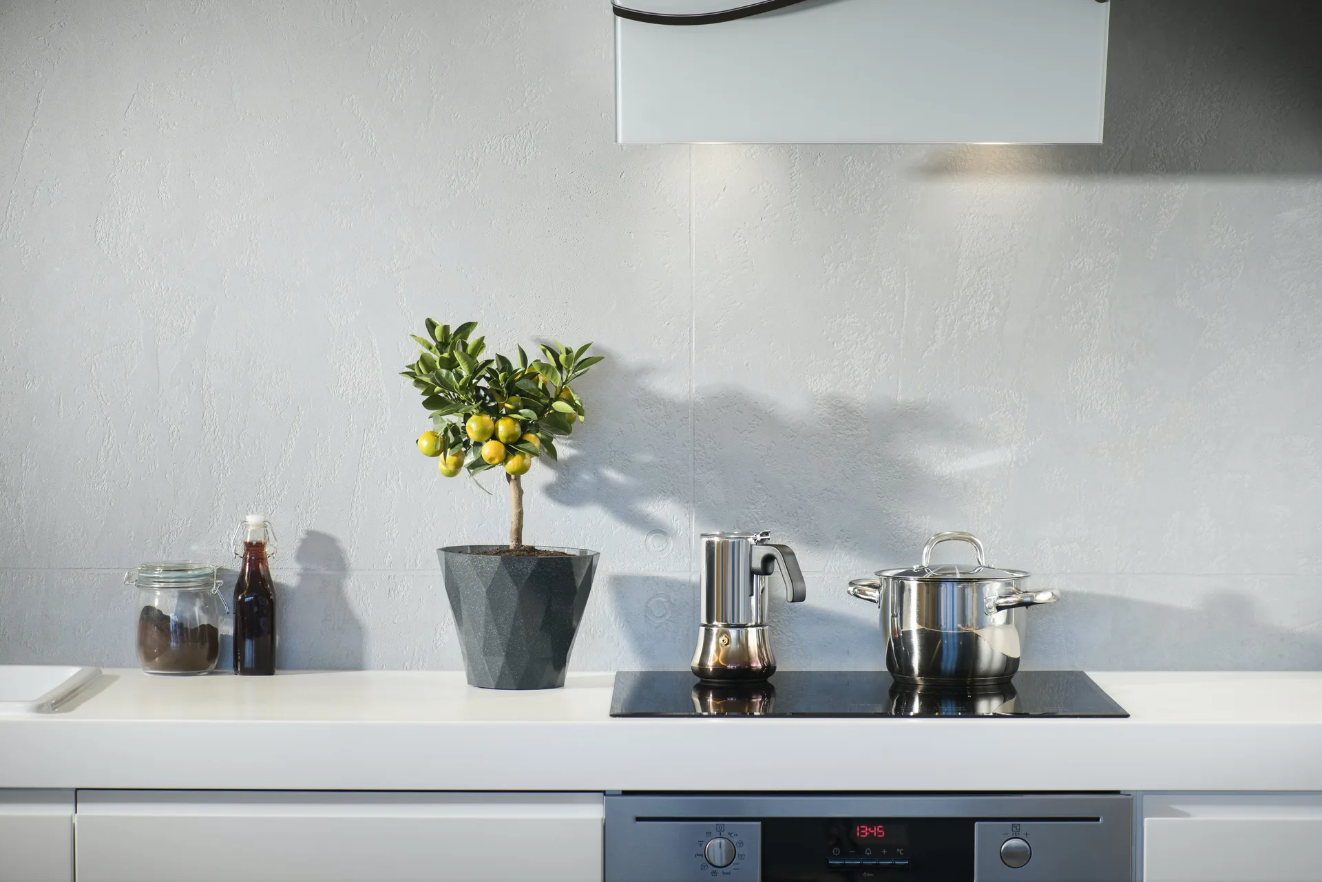 A modern kitchen counter features a sleek black cooktop, stainless steel cookware, a small potted citrus tree, and a light grey textured tiled backsplash.