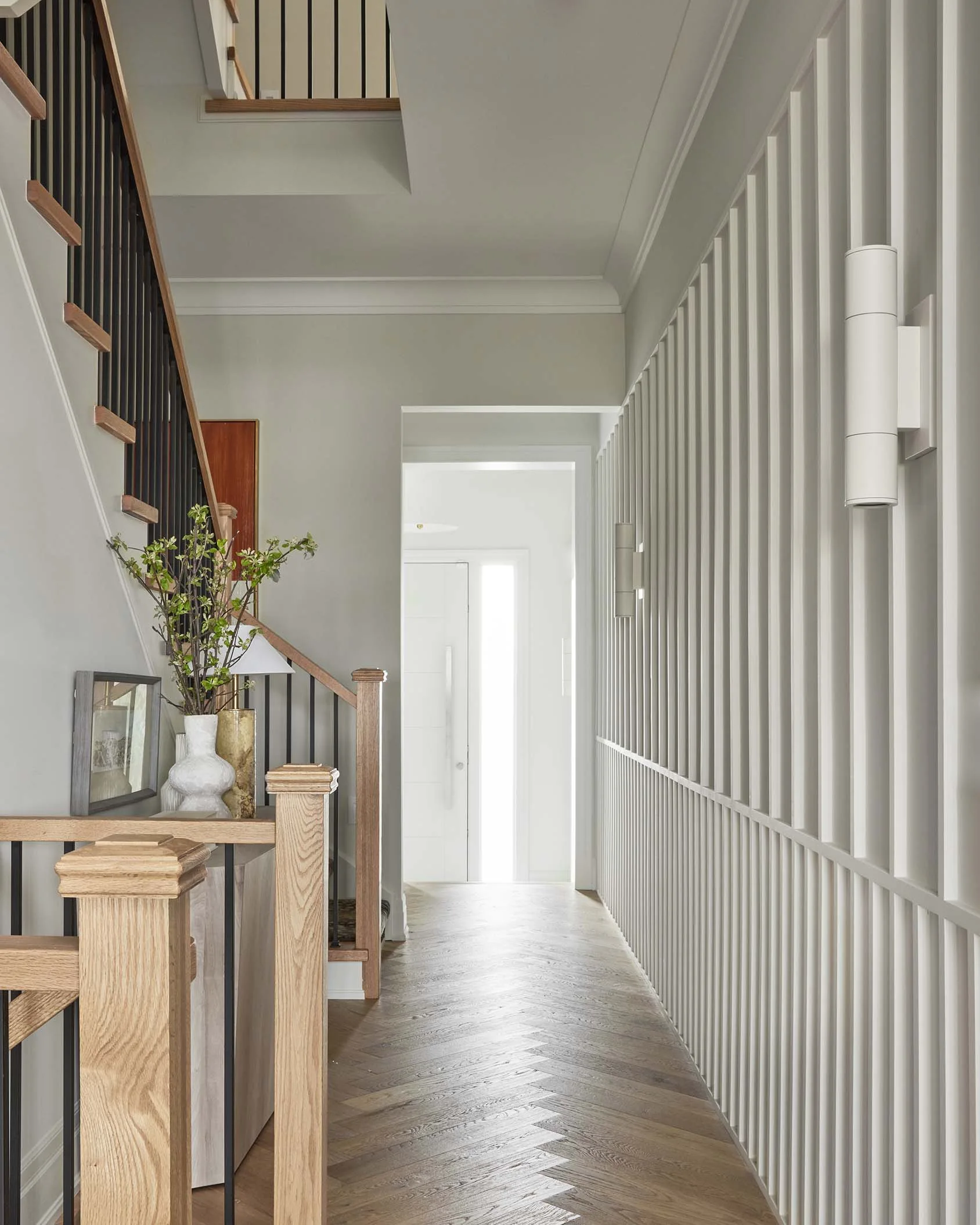 A hallway features a staircase on the left, a light gray slatted accent wall on the right, and light-toned herringbone hardwood flooring.
