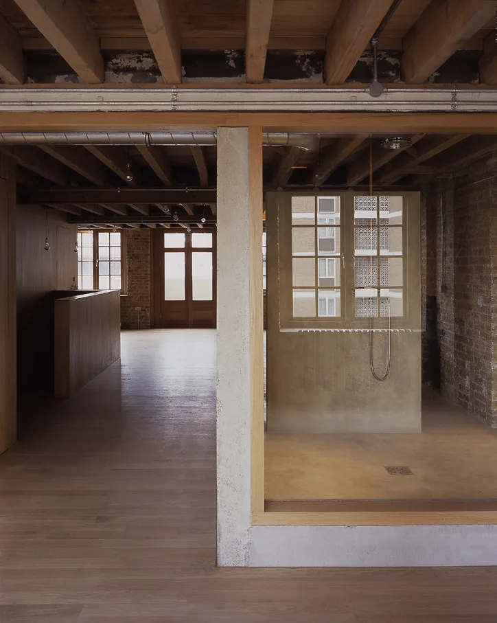 An industrial loft interior featuring a glass shower enclosure with concrete flooring, exposed timber ceiling joists, and brick walls.