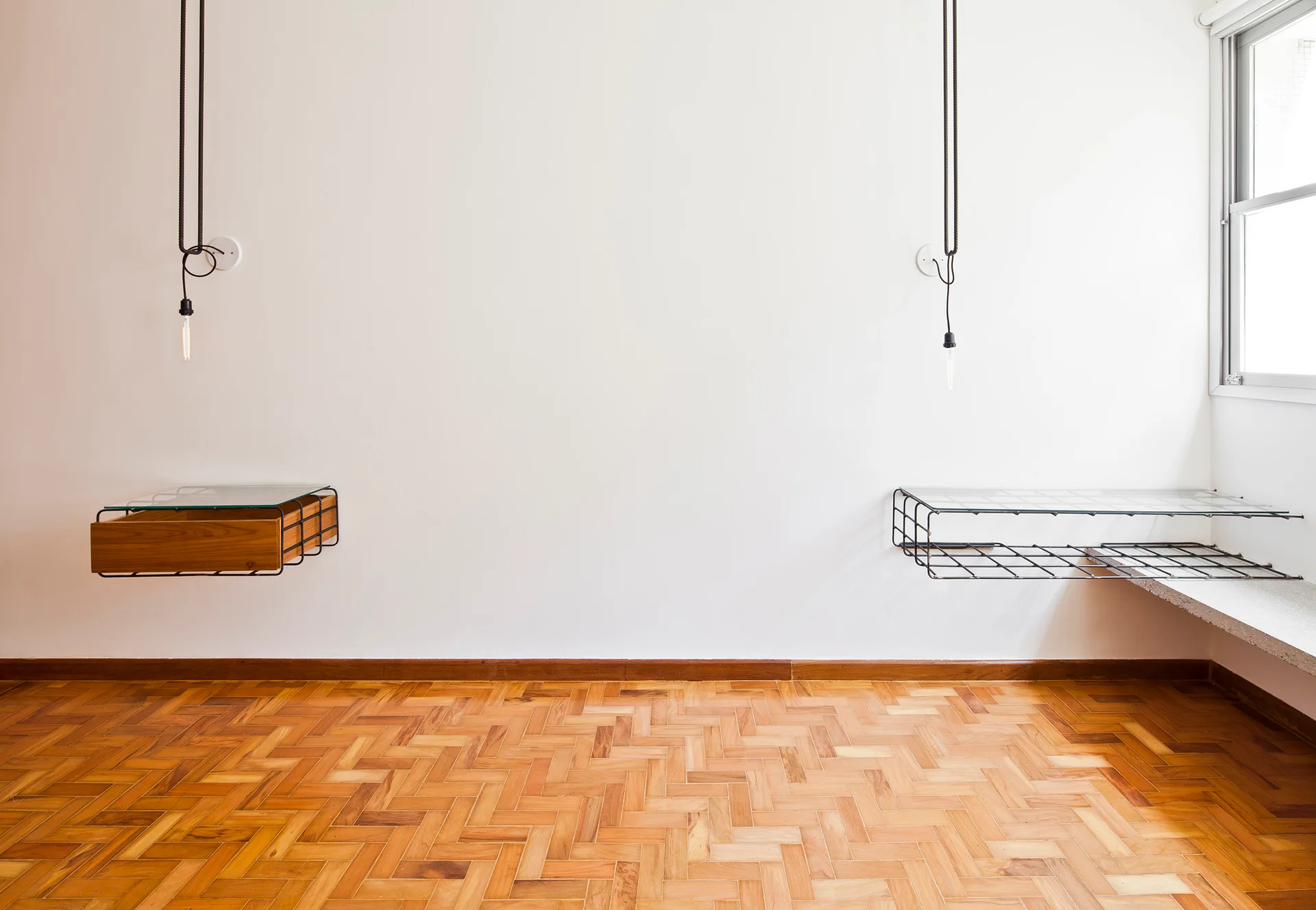 A white bedroom wall features two floating industrial bedside shelves of metal grid and wood under suspended cord bulb lights on parquet flooring.