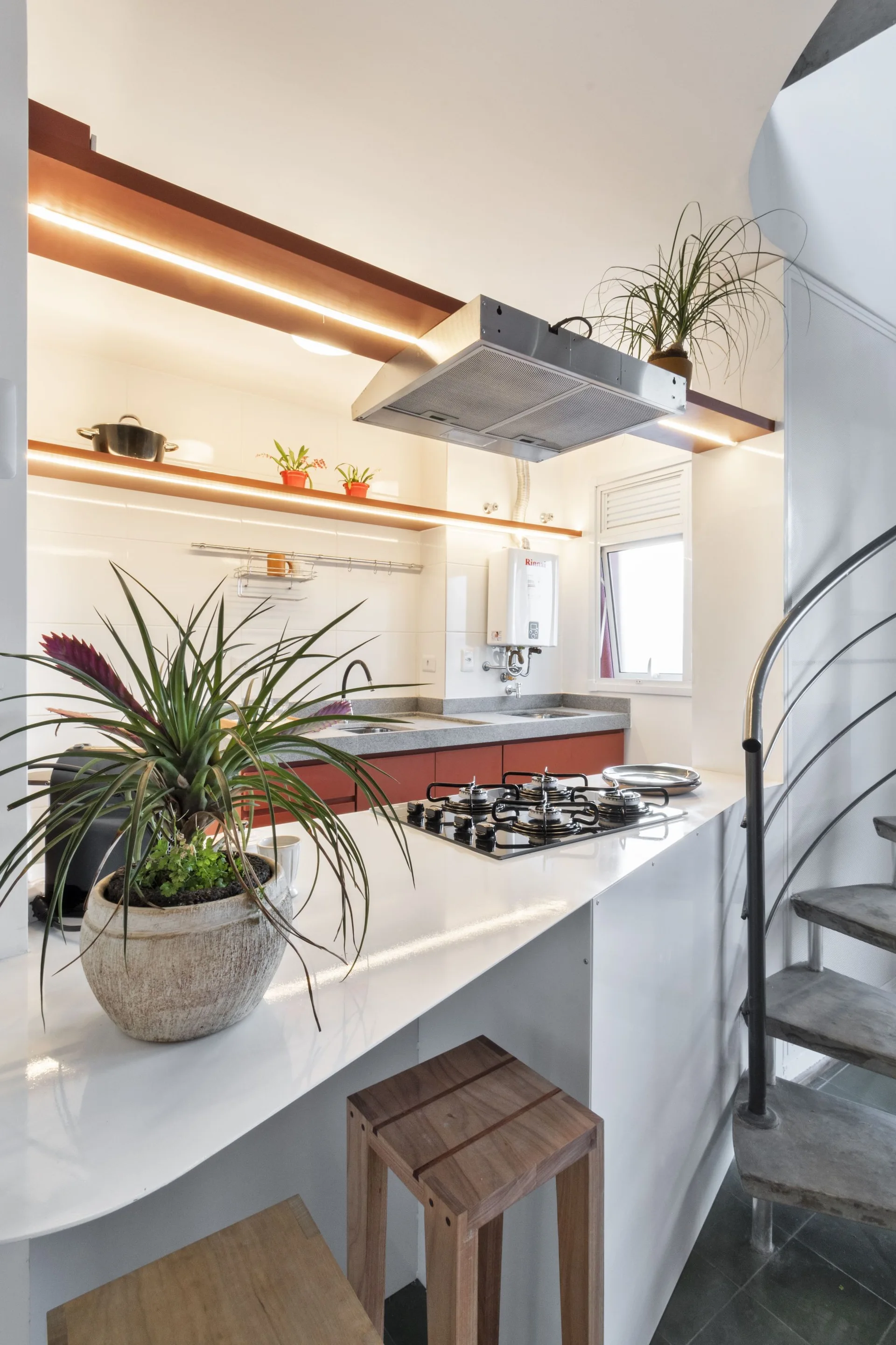 A compact kitchen features a white island with a gas cooktop, red lower cabinets, floating wooden shelves, and a metal spiral staircase.