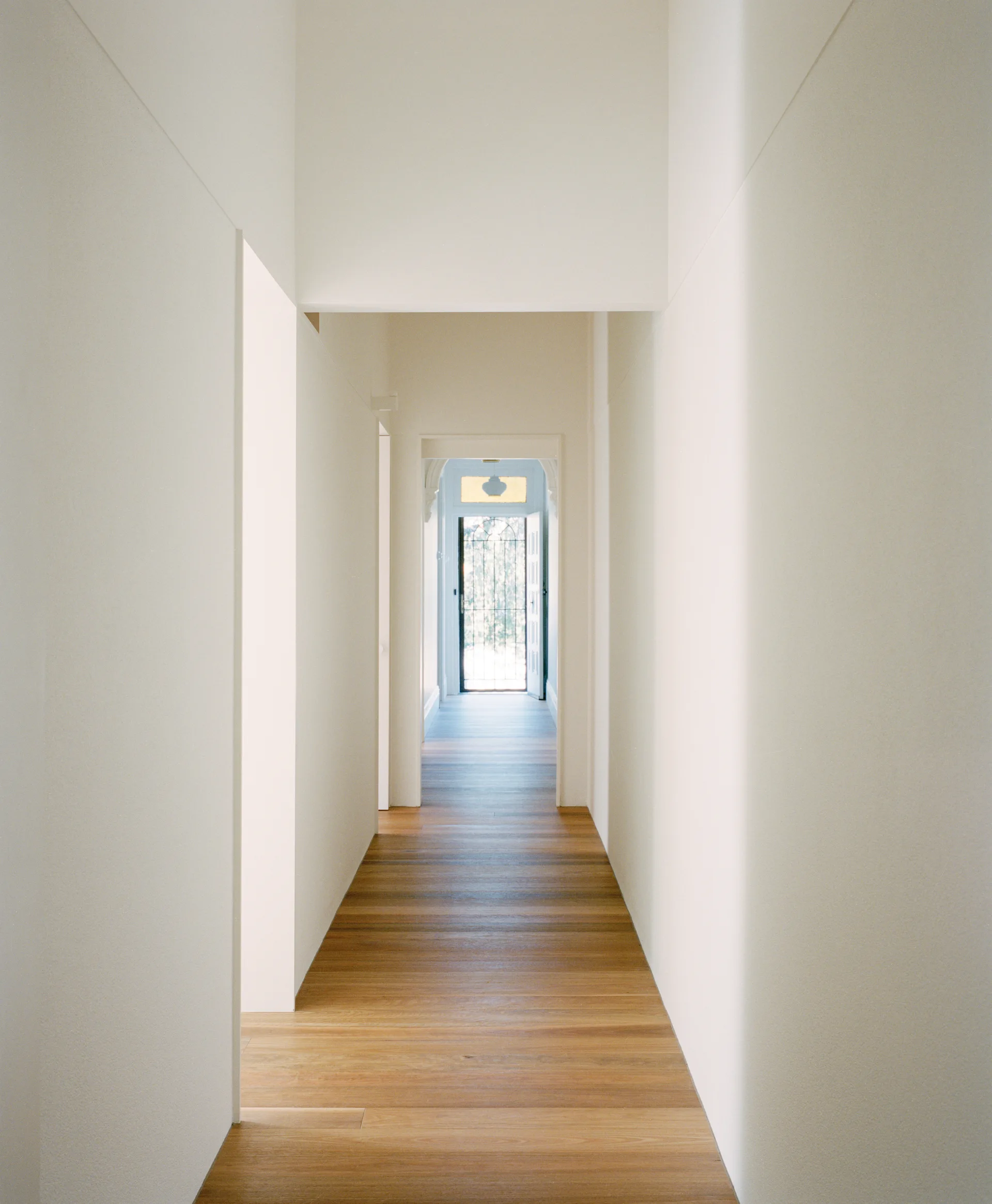 A long minimalist residential hallway with smooth white walls and warm wood plank flooring leads to an open doorway showing natural exterior light.