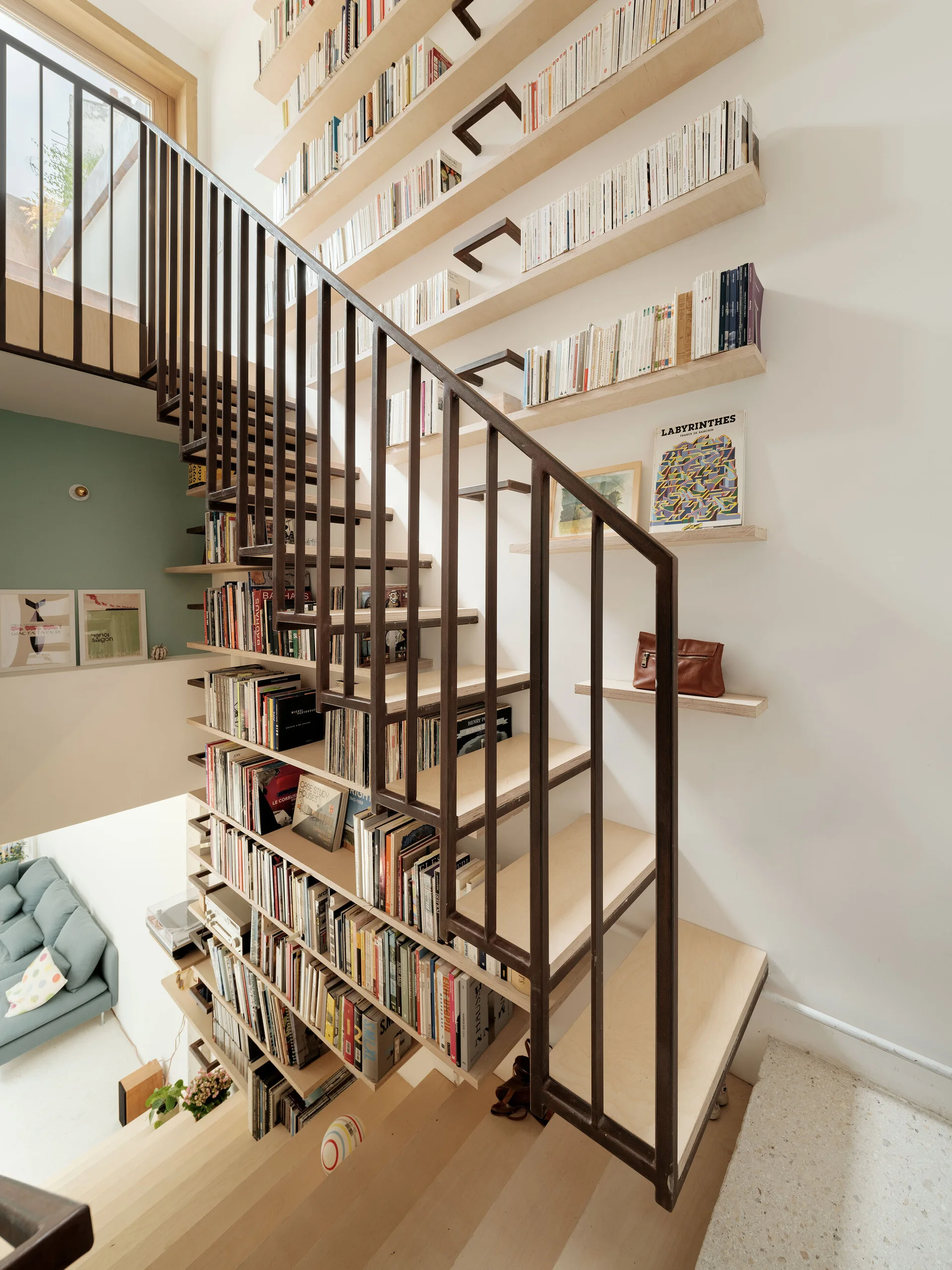 A light wood staircase with a dark metal railing features built-in floating bookshelves filled with books against a white wall.