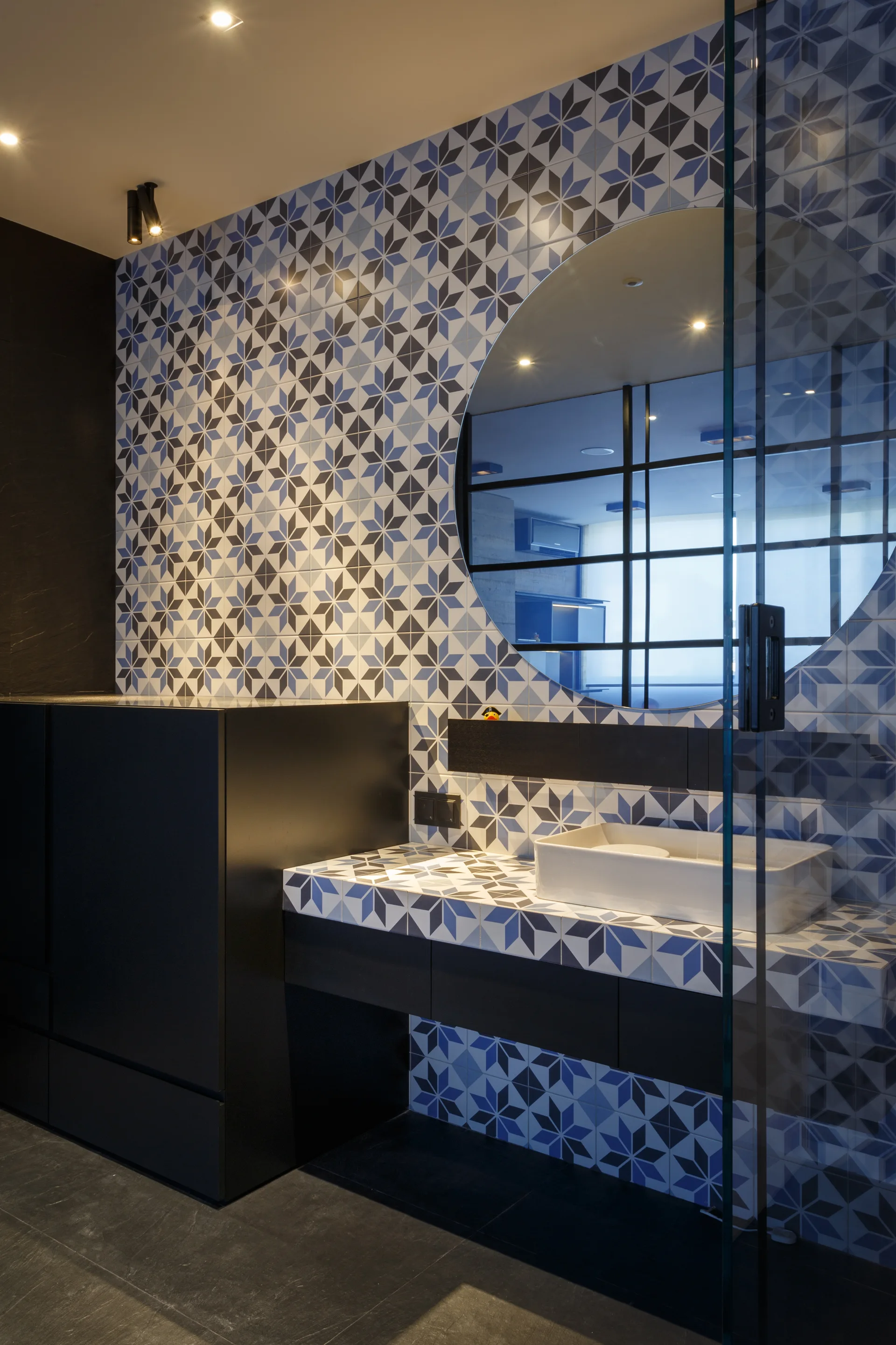 A bathroom vanity with a white vessel sink, matte black cabinets, and walls covered in blue and white geometric star tiles beside a round mirror.