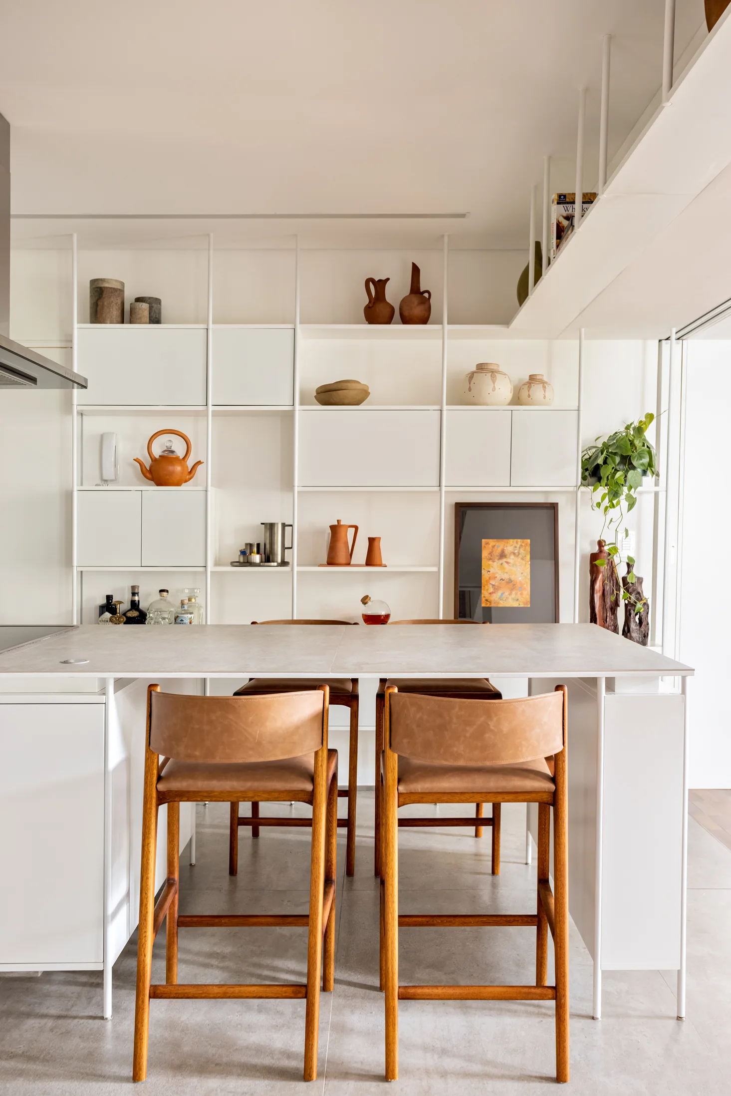 A white kitchen island with tan leather bar stools stands in front of a floor-to-ceiling white metal grid shelving unit holding ceramic pottery.