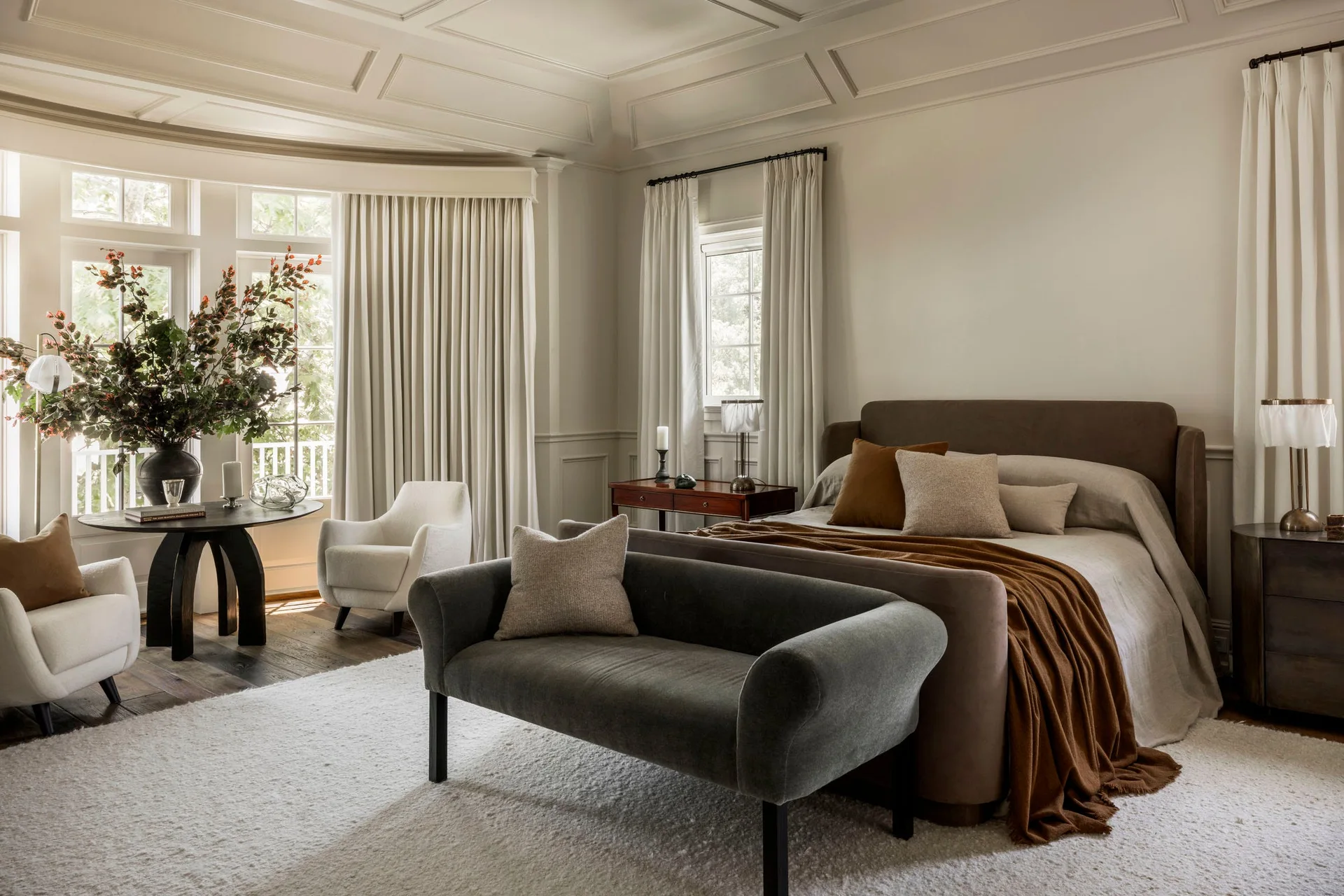 A bedroom featuring an upholstered brown bed, a charcoal gray bench, and off-white armchairs in a bay window area under a coffered ceiling.
