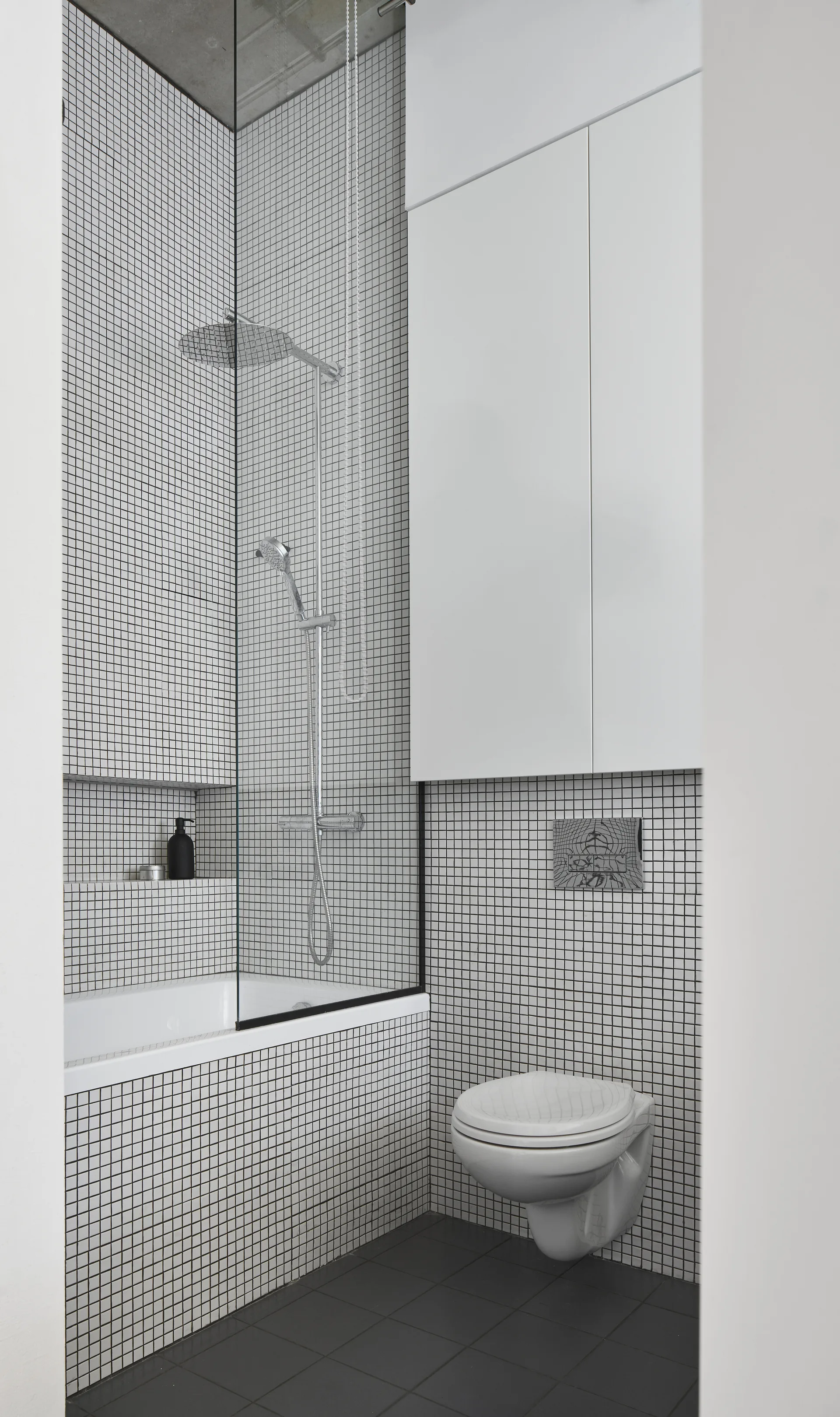 A minimalist bathroom features floor-to-ceiling white grid tiles, a wall-mounted toilet, a glass shower partition, and a sleek white upper cabinet.