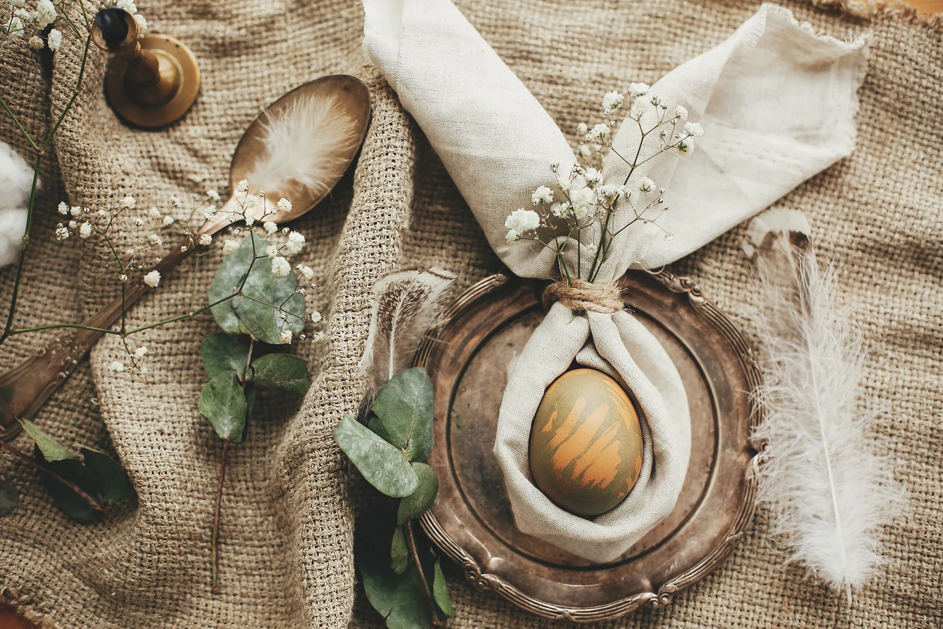 A top-down view of a rustic Easter table setting featuring a dyed egg wrapped in a bunny-ear linen napkin on a vintage metal plate.