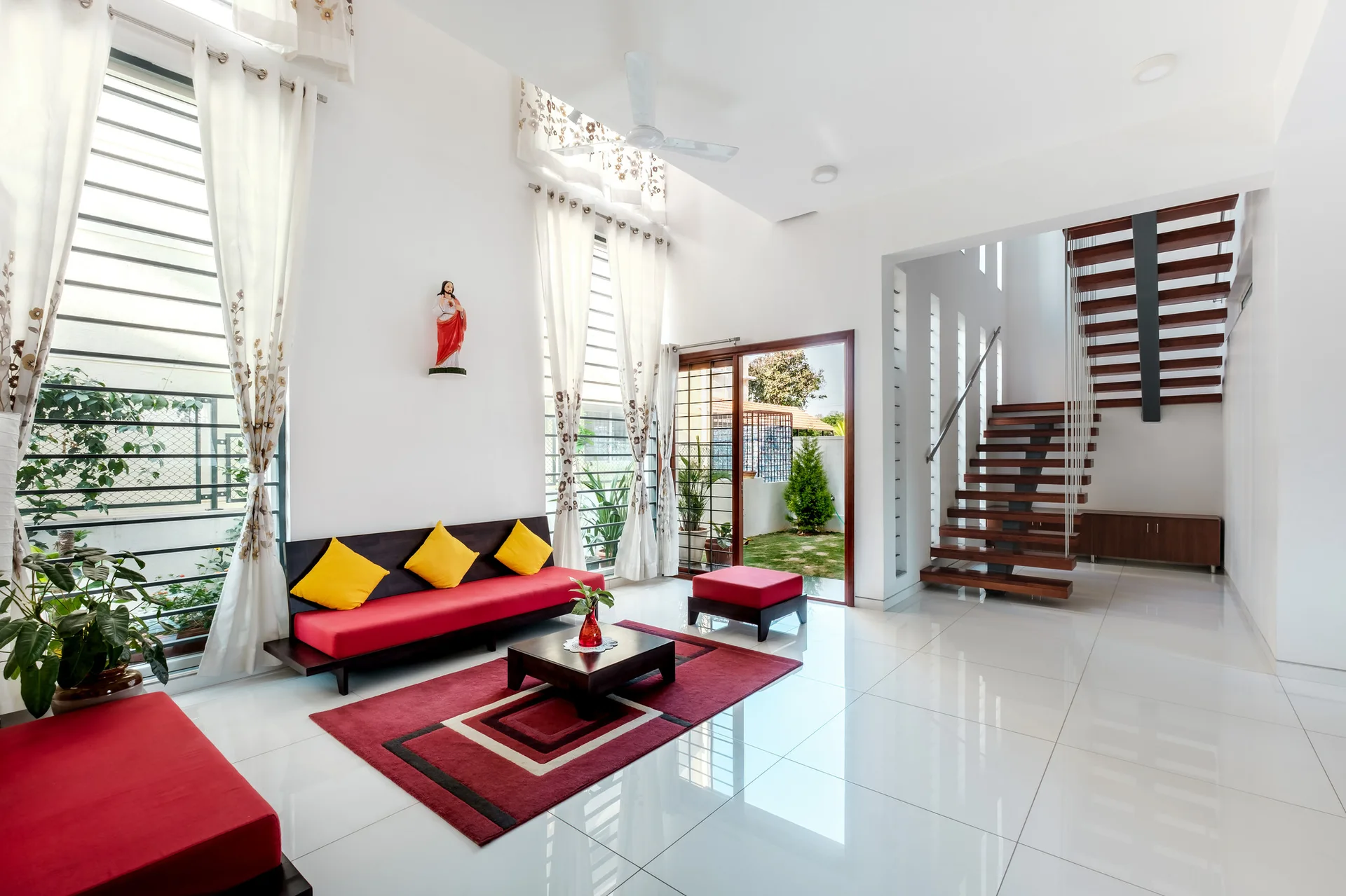 A bright living room features a red sofa with yellow cushions on a glossy white tile floor near a floating wooden staircase.