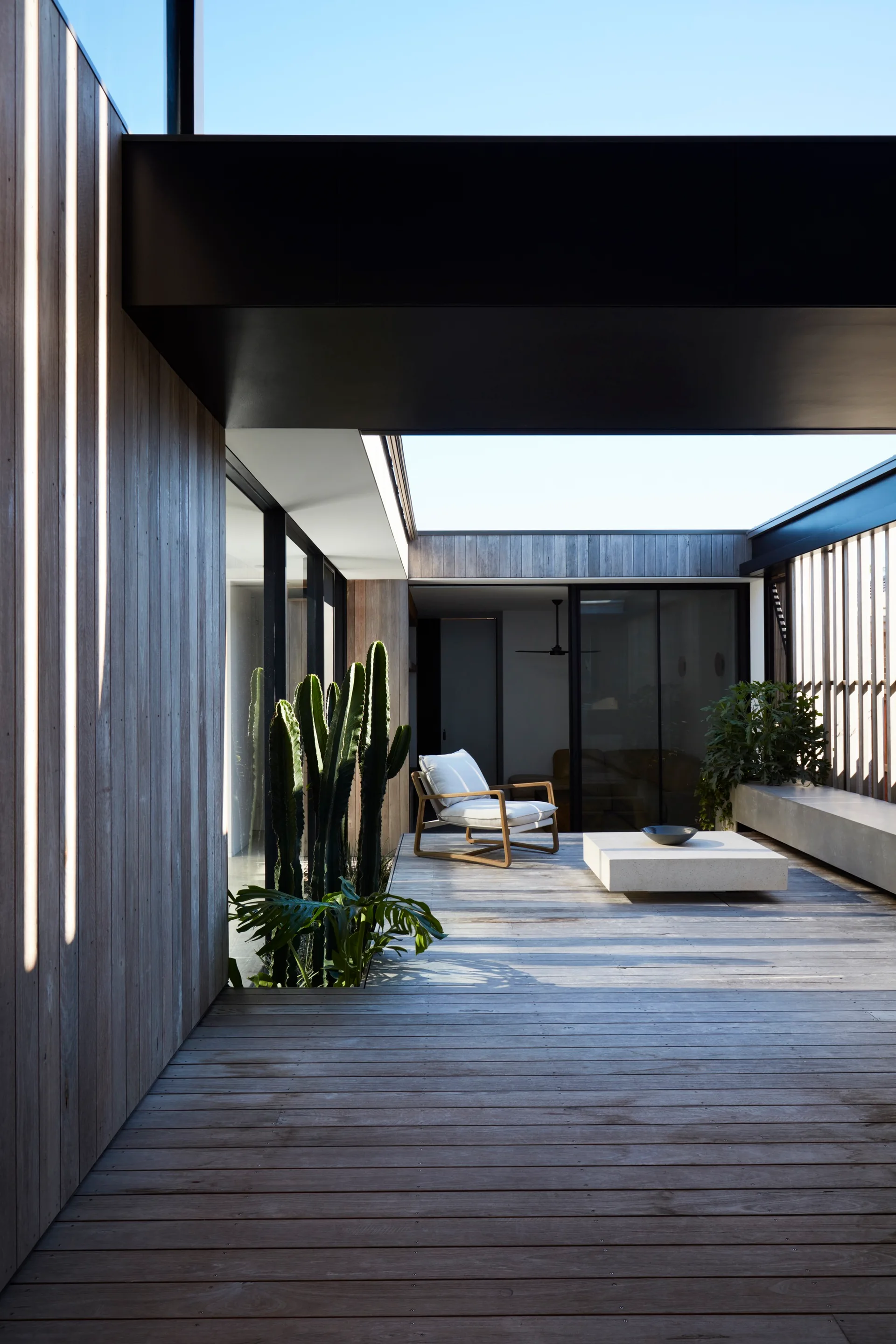 A minimalist outdoor courtyard featuring a light wood deck, a modern white lounge chair, a low square stone coffee table, and a tall green cactus.