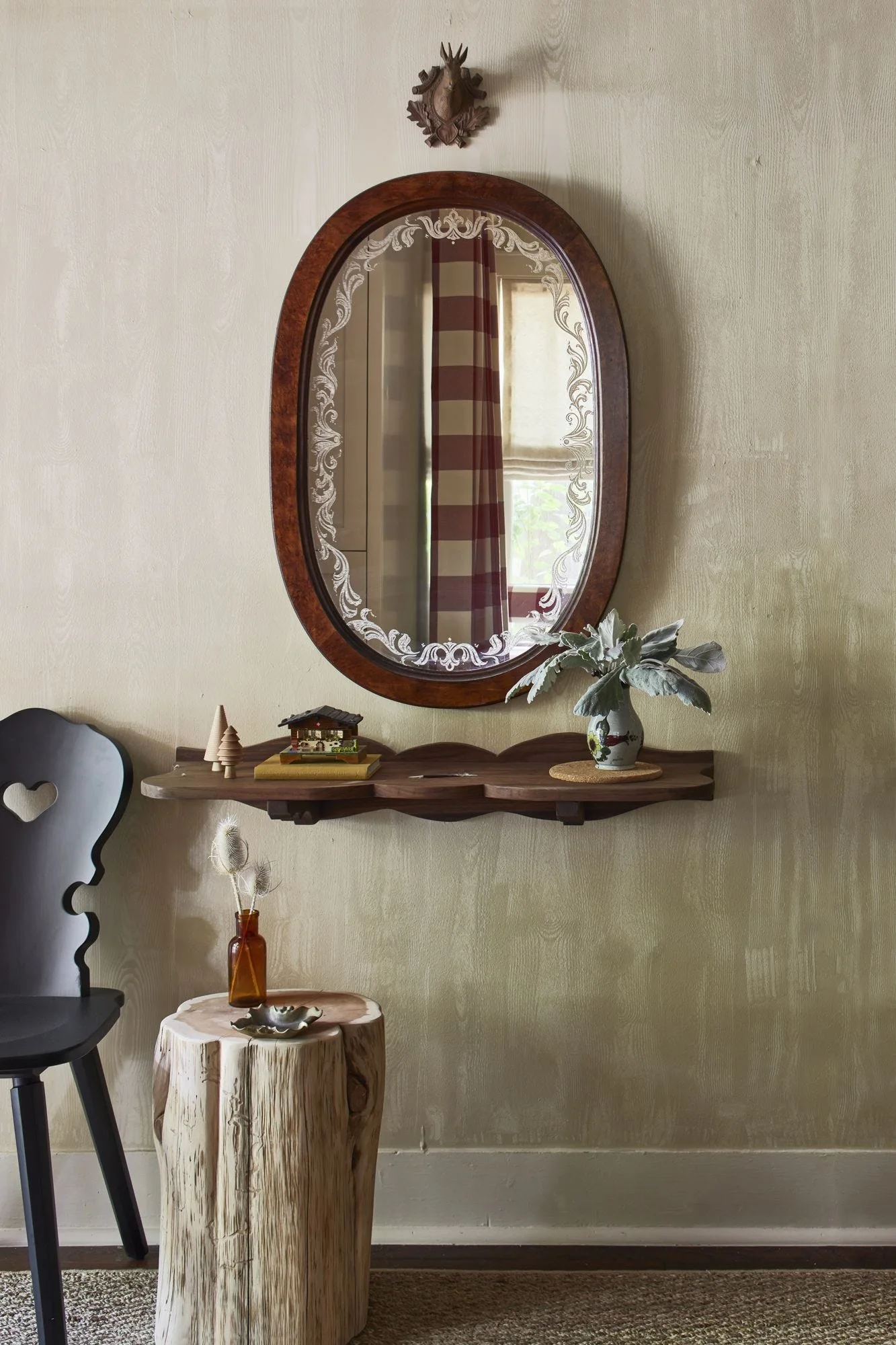 An entryway vignette features an oval etched mirror above a scalloped wood shelf, flanked by a black heart-back chair and a tree stump table.