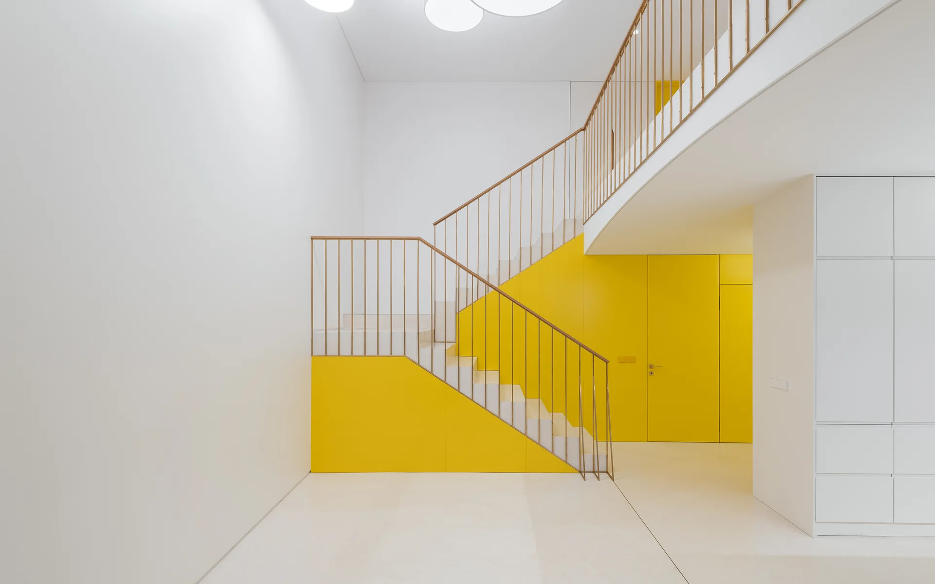 A minimalist staircase features light stone steps, a slim vertical metal railing, and a vibrant yellow accent wall with a flush integrated door.