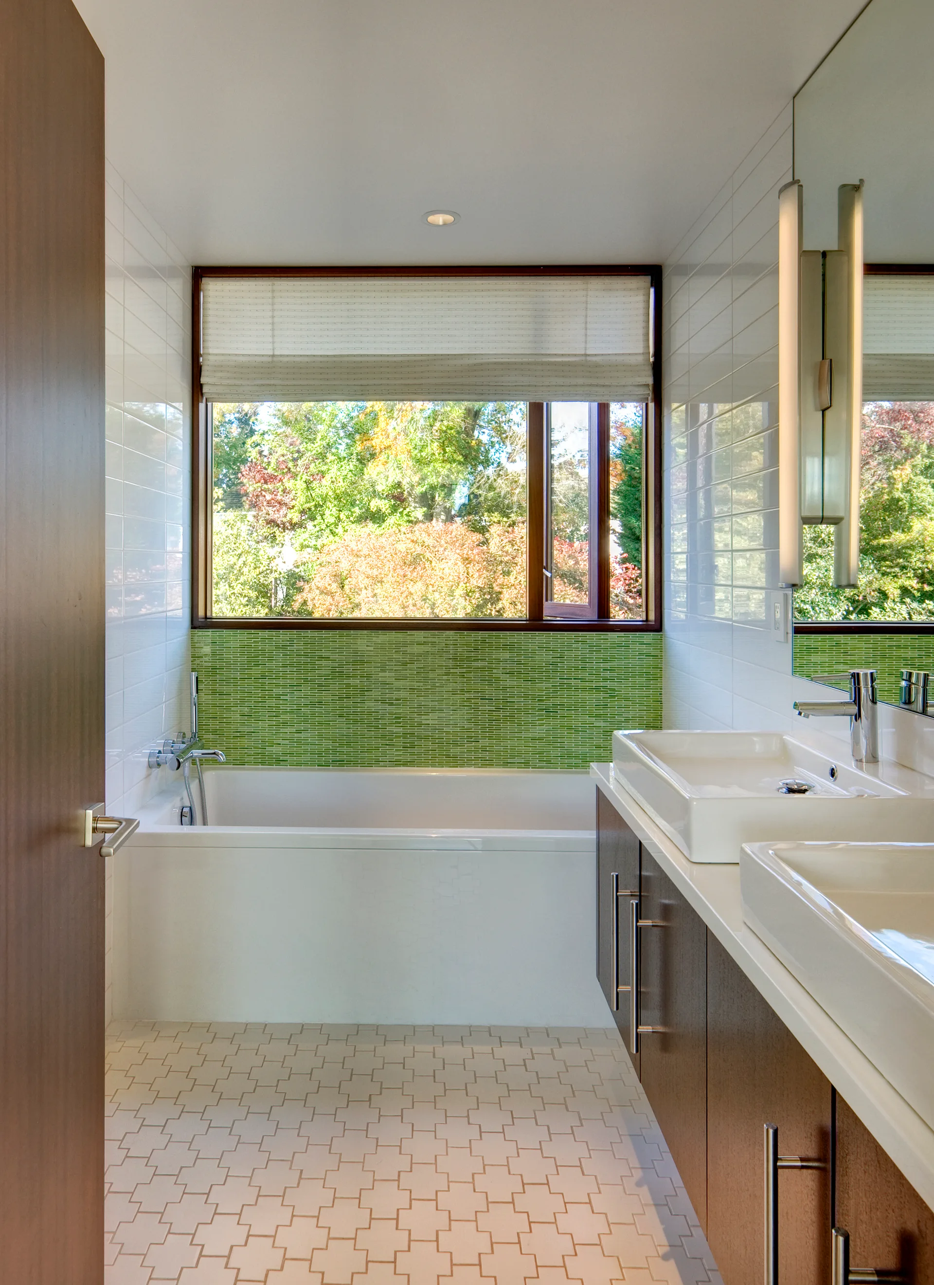 A modern bathroom featuring a white built-in bathtub below a window, a green mosaic tile backsplash, and a double wood vanity with vessel sinks.