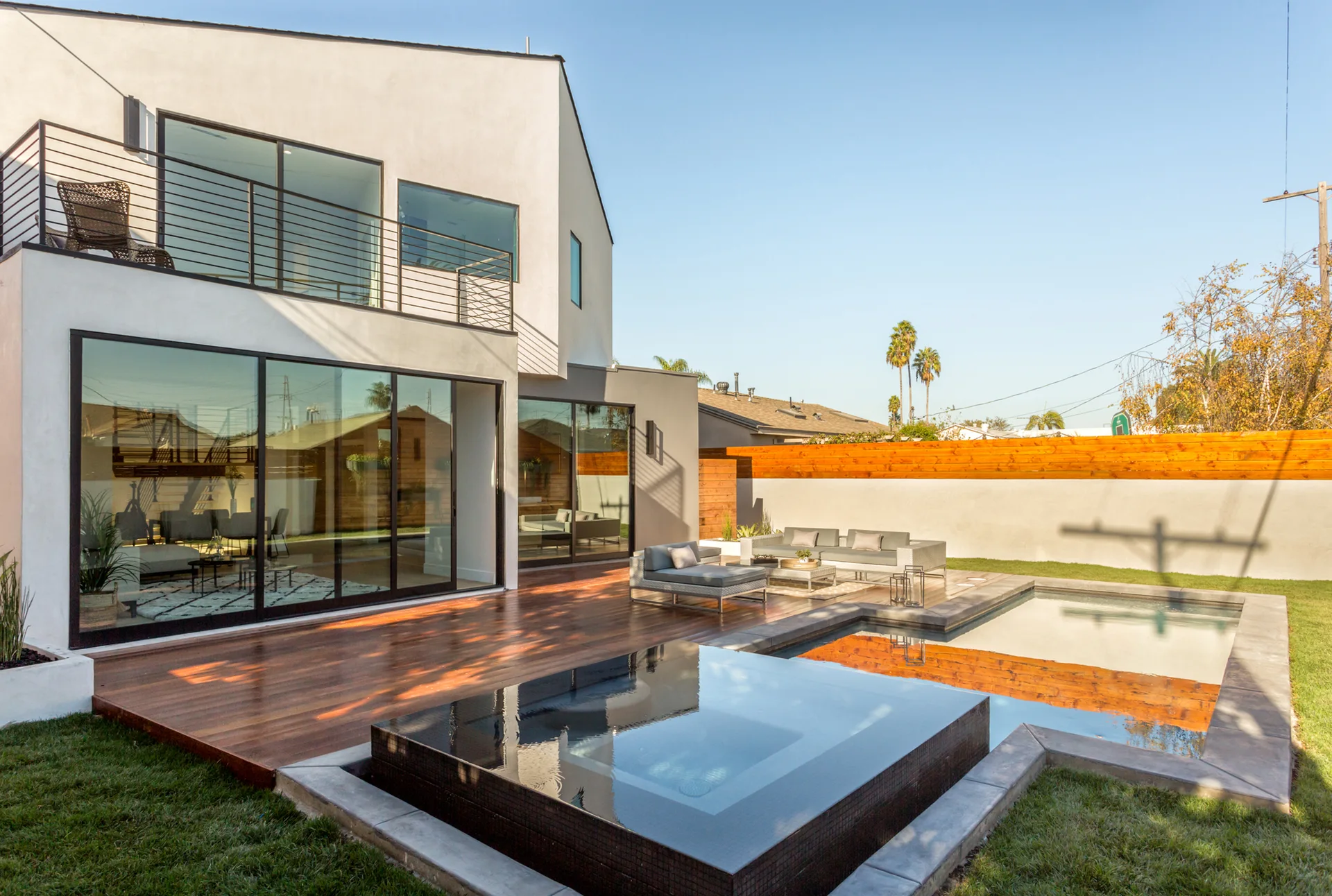 A modern two-story white stucco home features a wooden deck patio, grey outdoor sofa, and a swimming pool with an integrated dark-tiled spa.