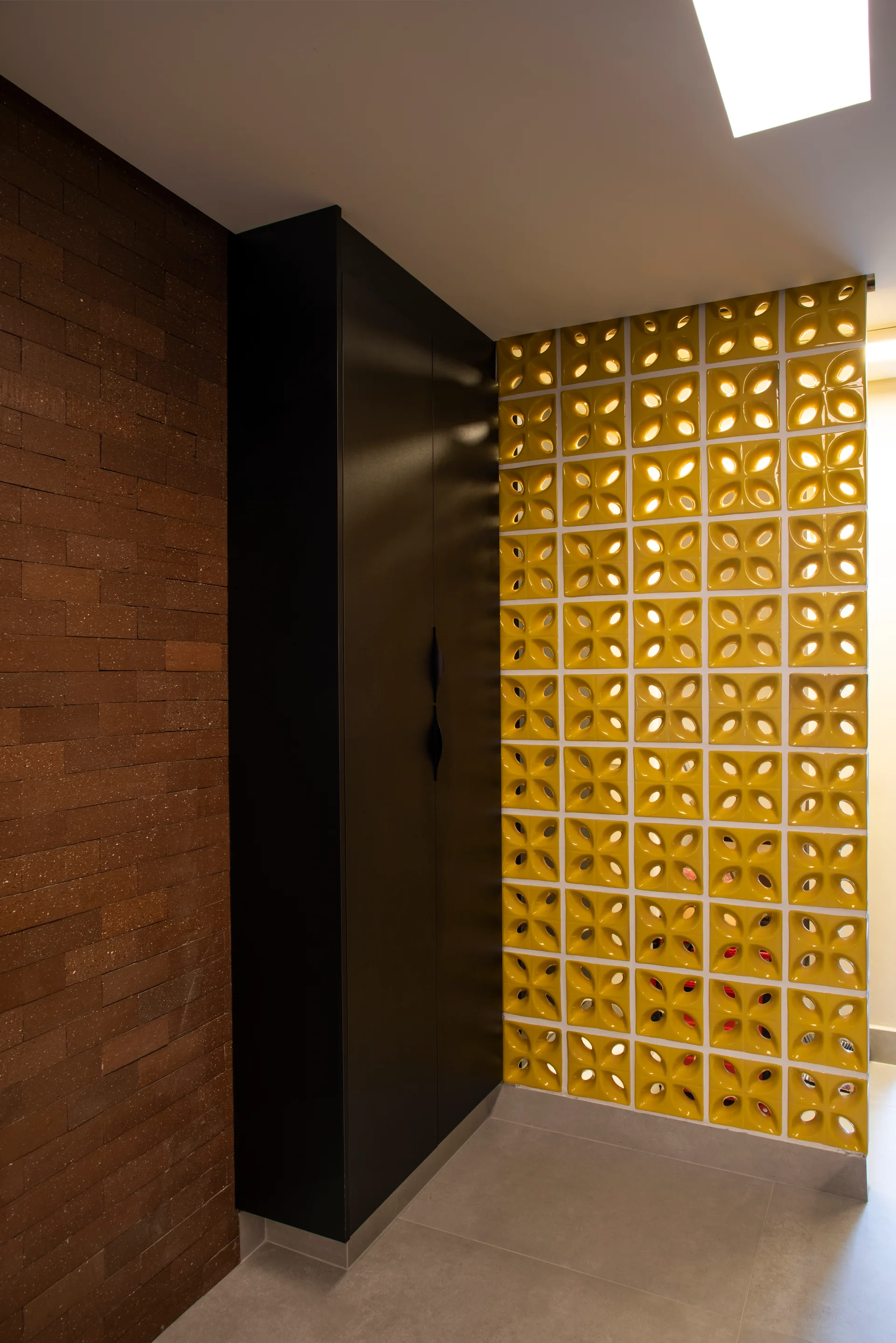 A tall black cabinet built in between an exposed brown brick wall and a yellow geometric breeze block partition over grey tiles.