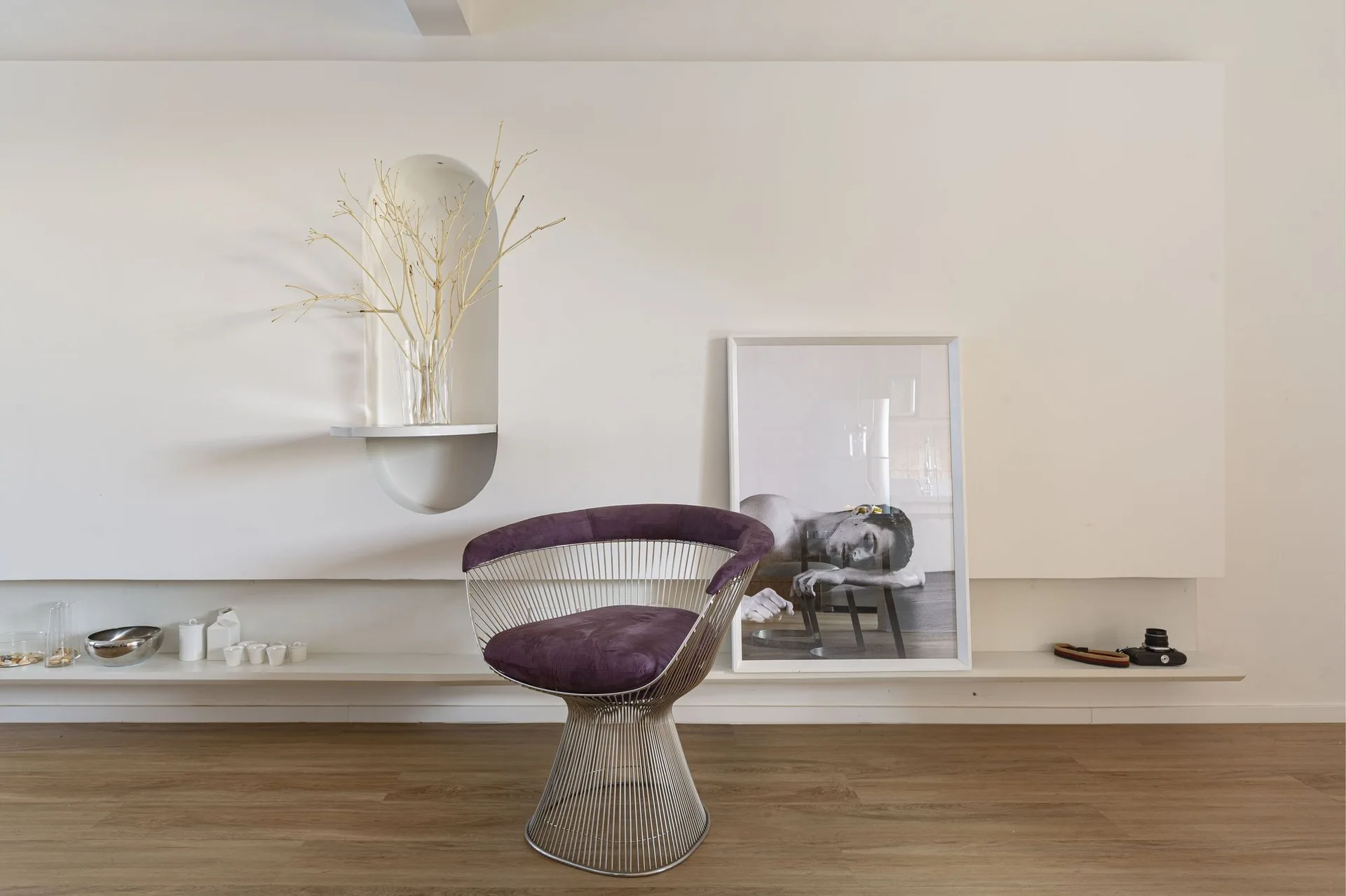 A purple wire pedestal armchair sits on wood flooring in front of a white wall with an arched recess and a low display shelf.