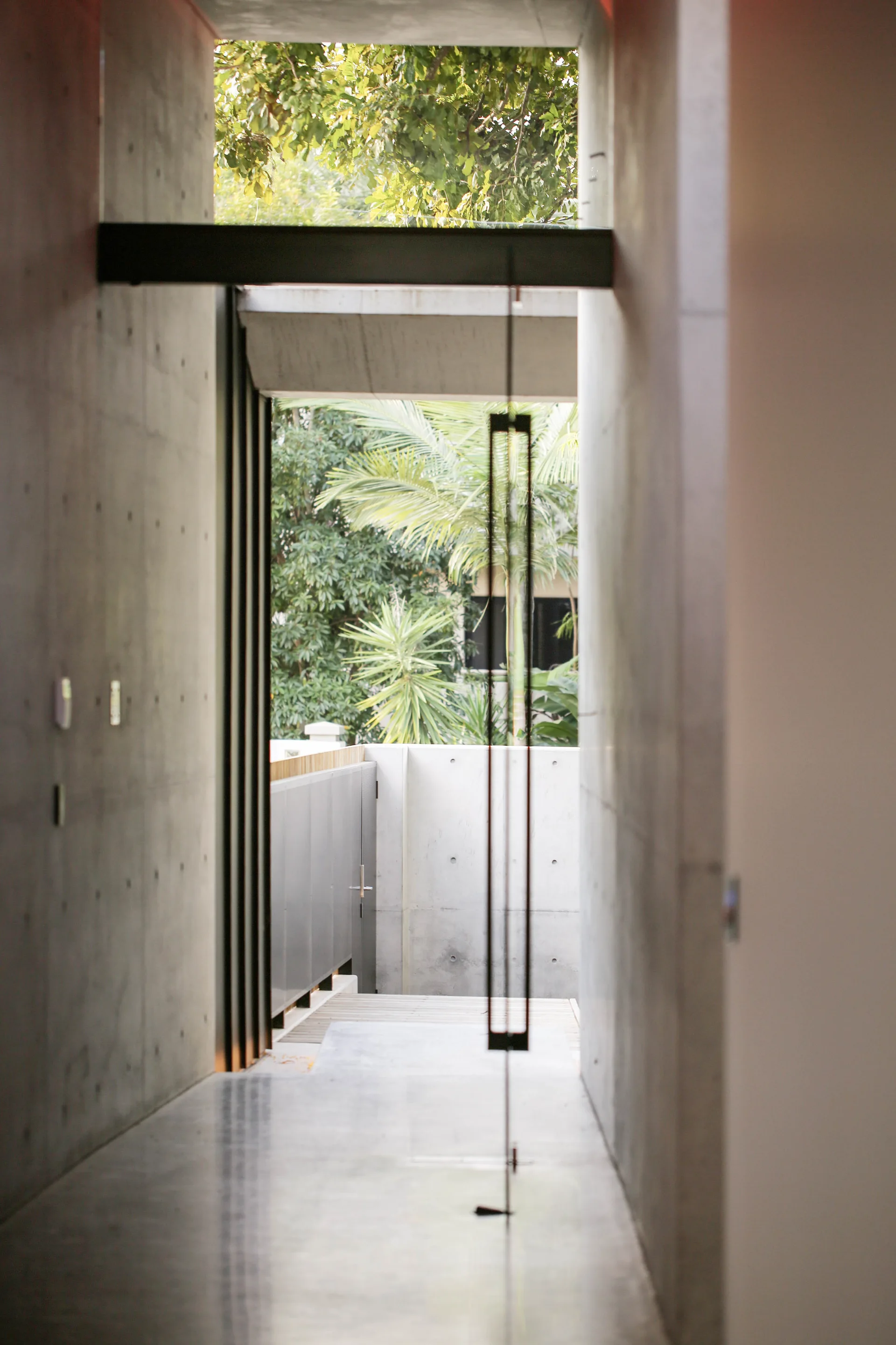 A long minimalist hallway features cast concrete walls with formwork tie marks and a tall metal-framed glass pivot door opening to an outdoor garden.