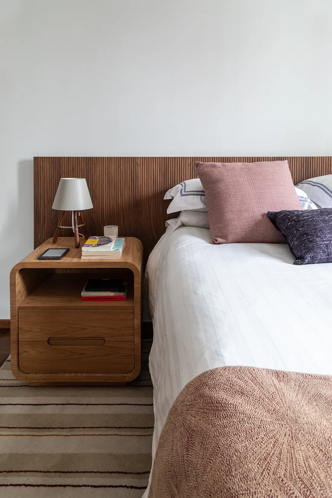 A bedroom featuring a bed with white and pink bedding, a fluted wood headboard, a curved wooden nightstand with a lamp, and a striped rug.