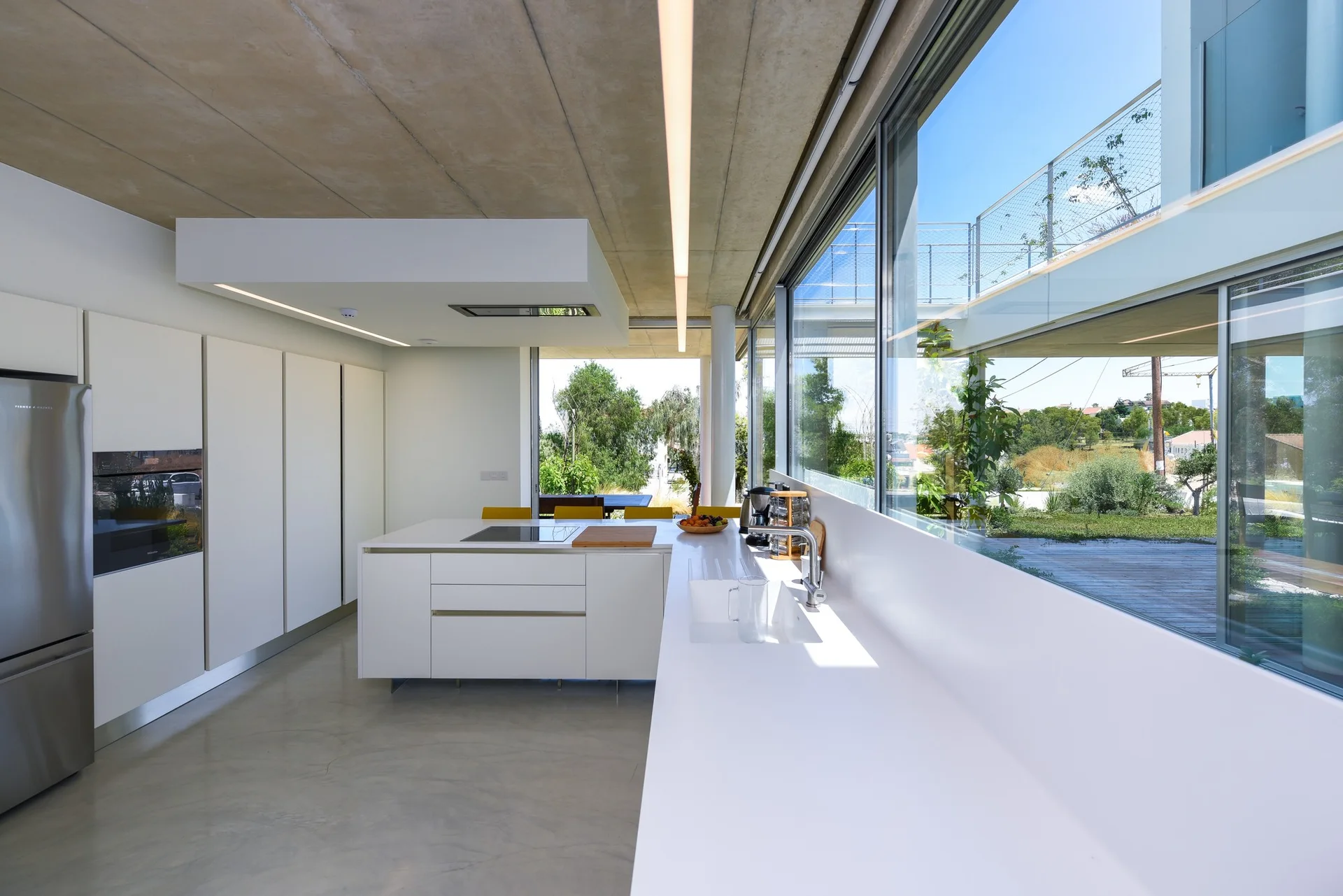 A modern white kitchen features seamless cabinetry, a central island, a concrete ceiling, and a wide horizontal window showing an outdoor landscape.