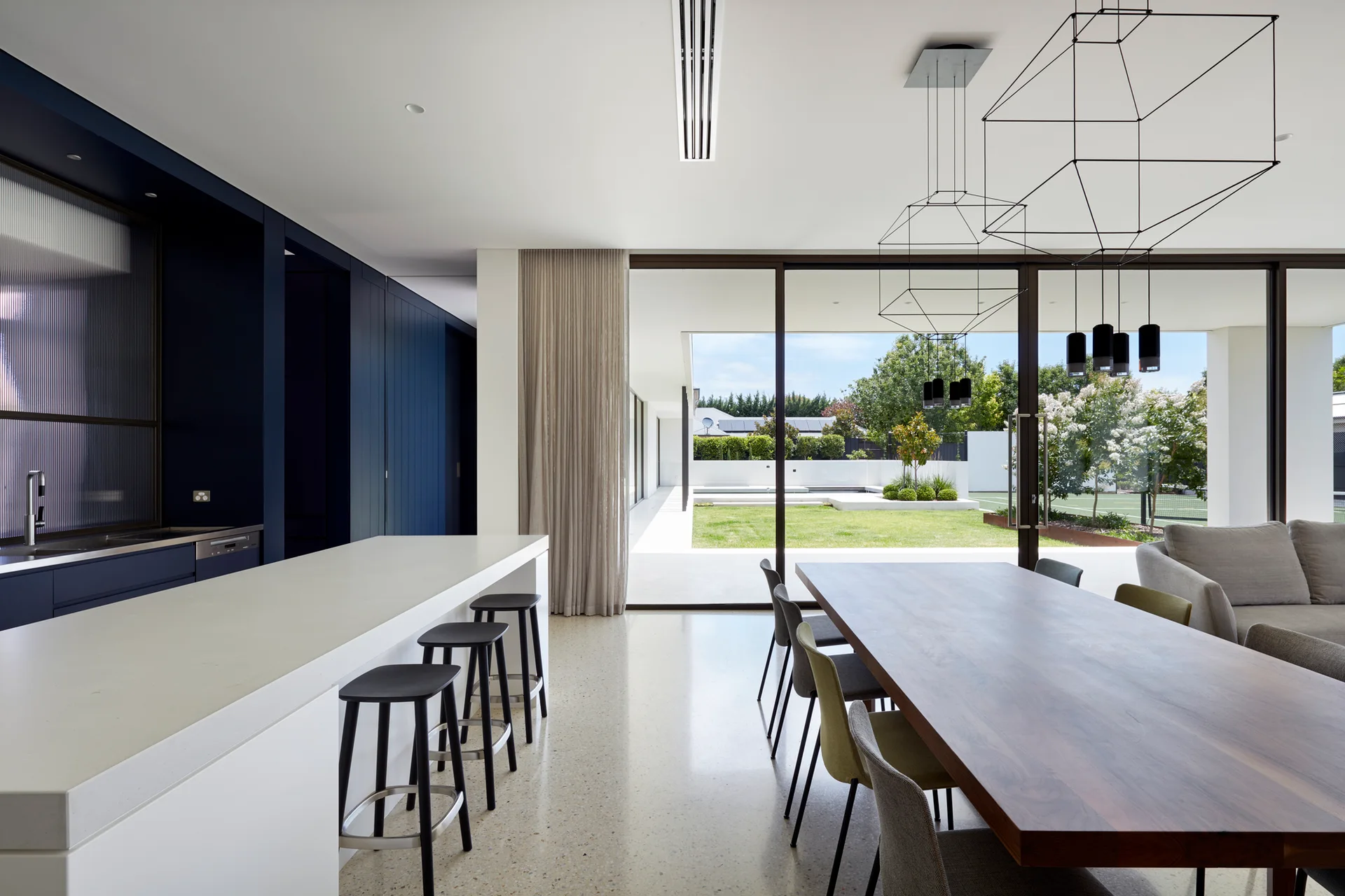A modern kitchen and dining area with dark blue cabinets, a white island, and a wooden table near large glass doors opening to a yard.