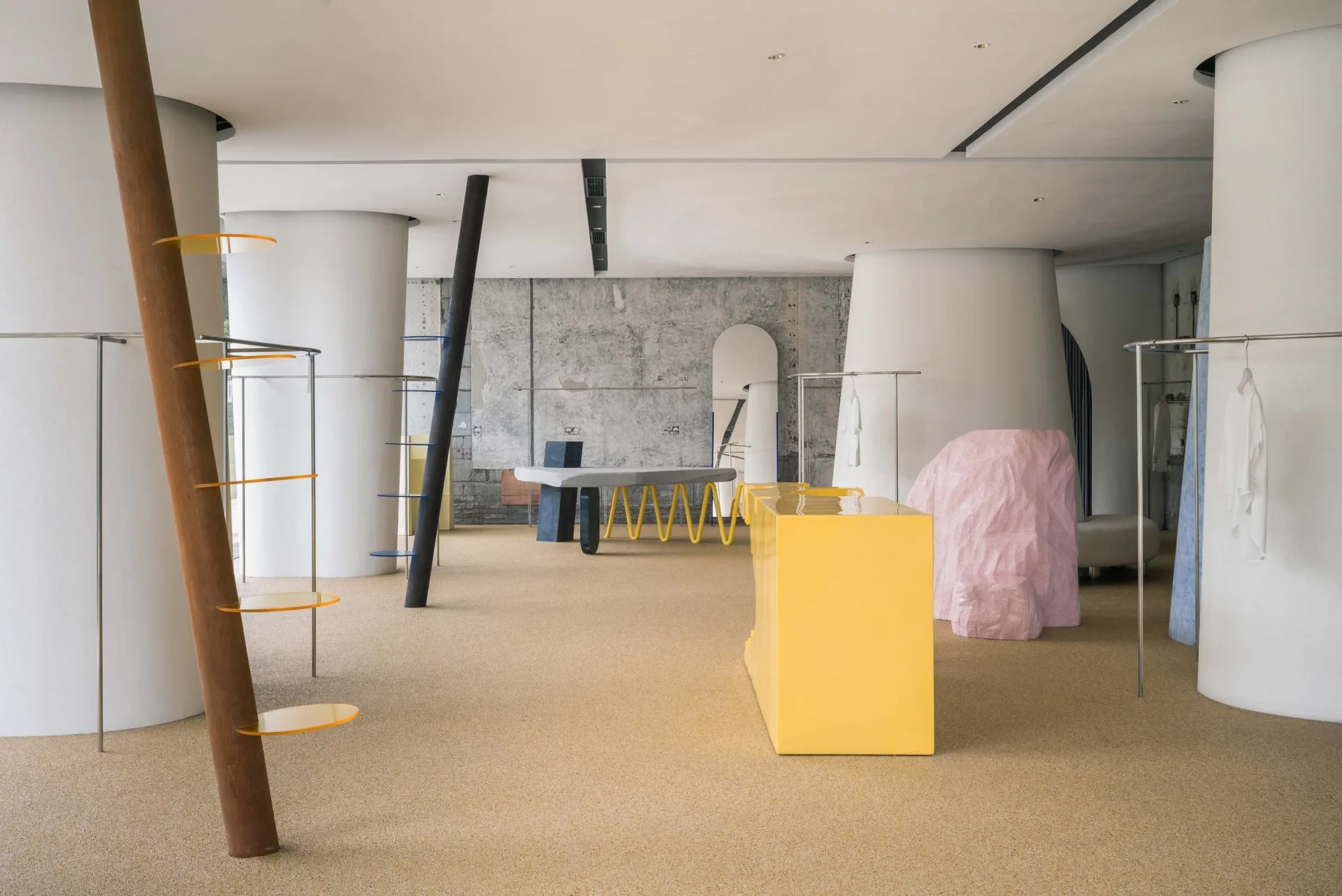 A modern retail showroom features white columns, a glossy yellow display counter, a textured pink sculpture, and clothing racks on a speckled floor.