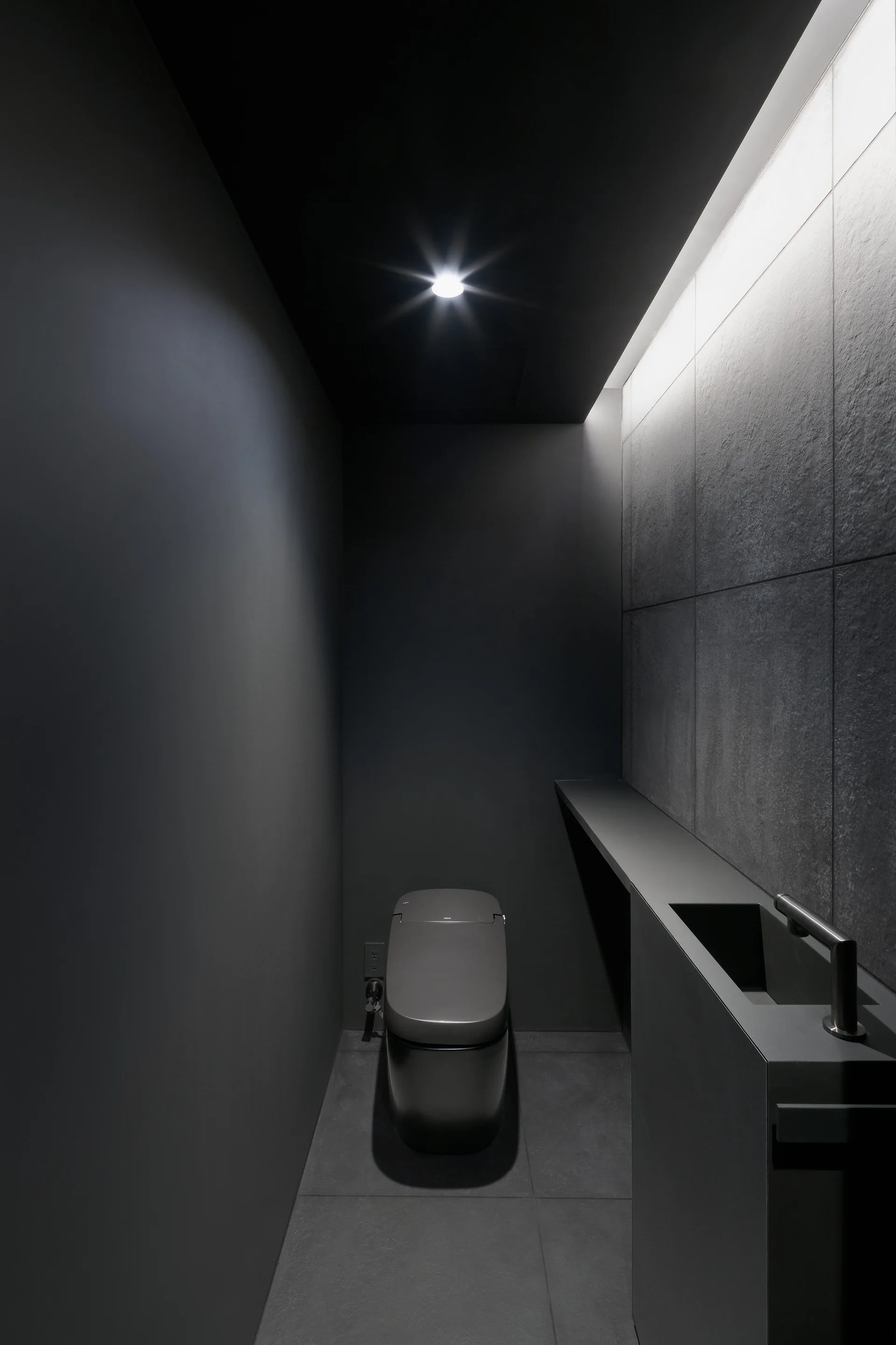 A narrow minimalist powder room in all-black featuring a modern matte toilet, an integrated console sink, and a textured charcoal tiled wall.
