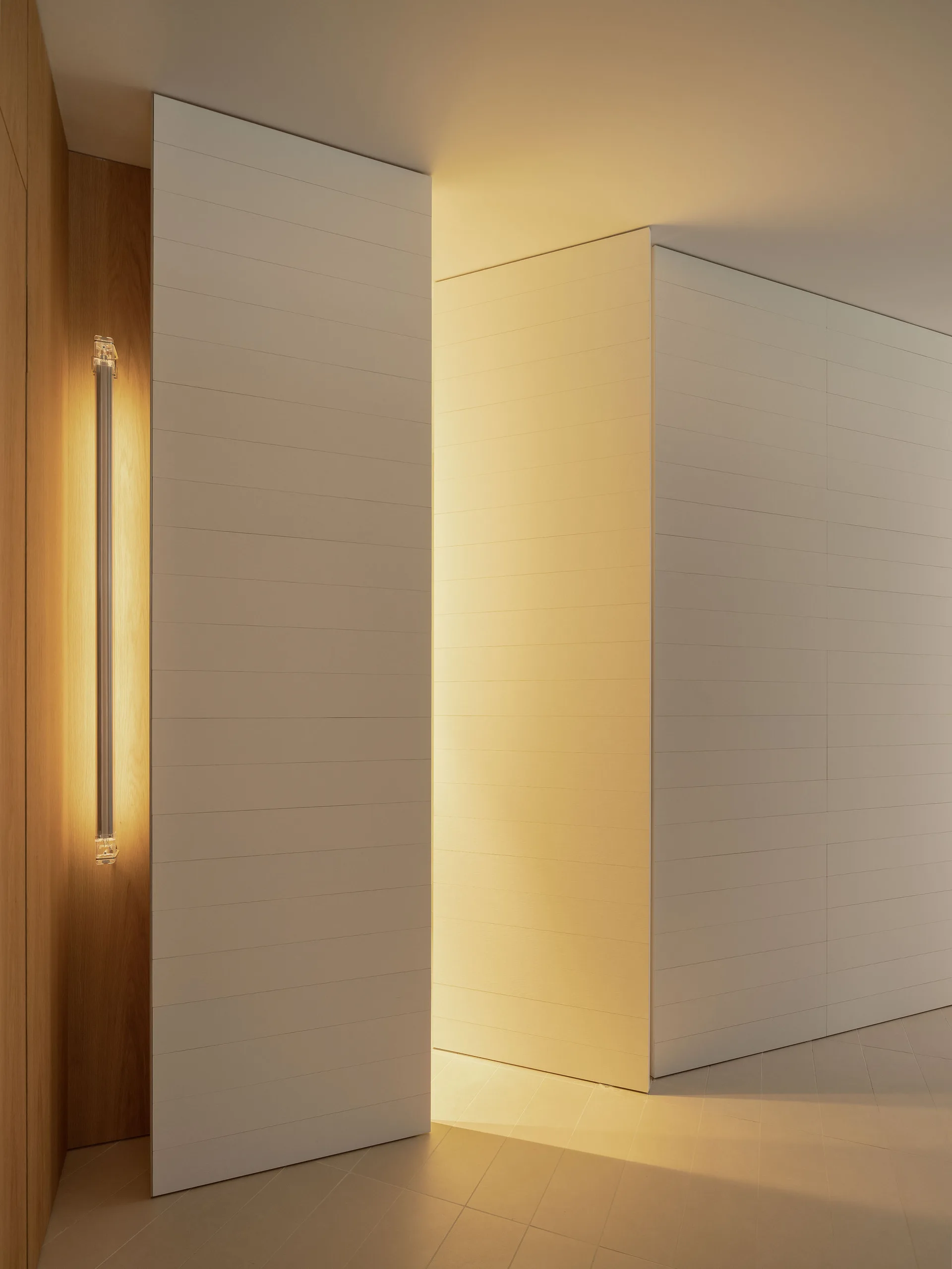 White horizontal-planked wall panels feature an integrated pivot door with warm golden light glowing from behind, next to a vertical glass wall sconce.