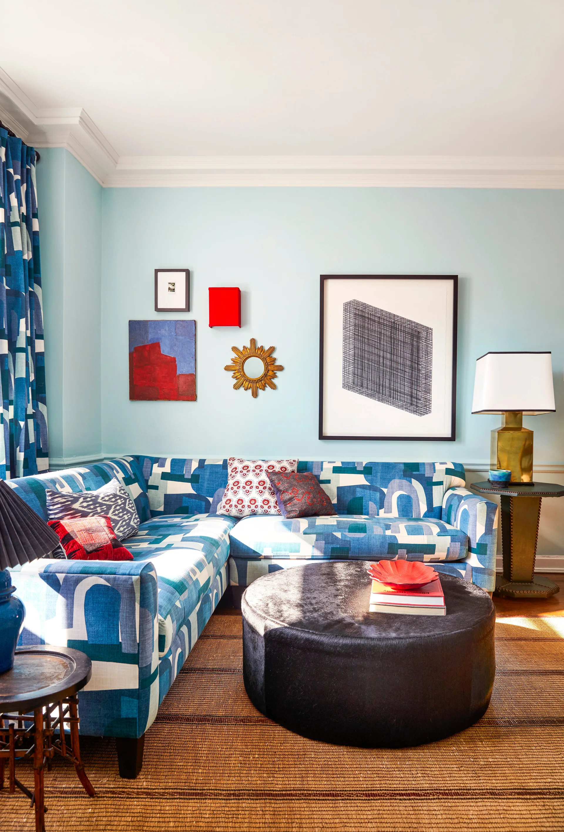 An eclectic living room featuring a patterned blue sectional sofa, a round black ottoman coffee table, and a small gallery wall on a light blue wall.