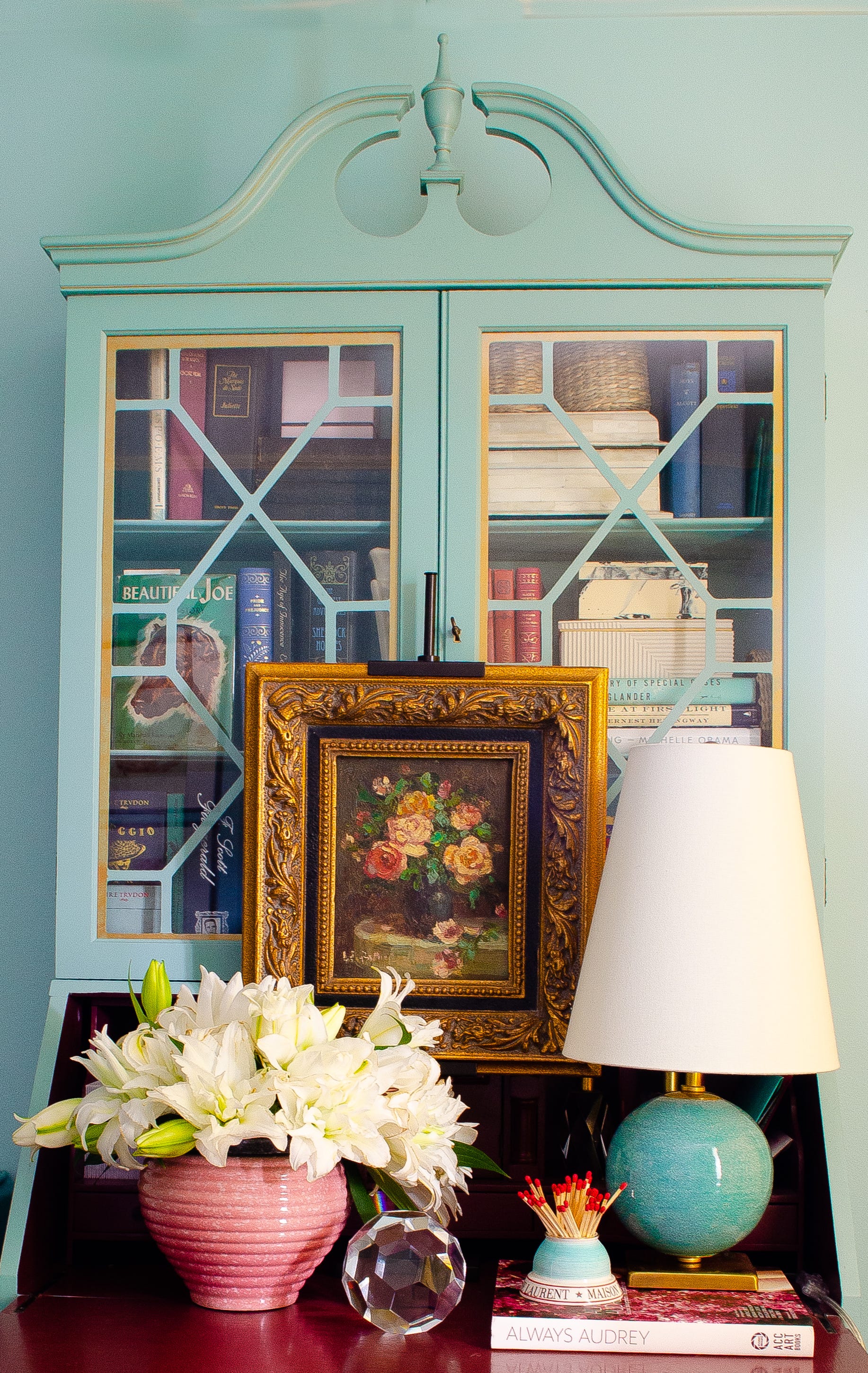 An aqua blue painted secretary desk with lattice glass doors displays an ornate gold-framed floral painting, a ceramic table lamp, and white lilies.