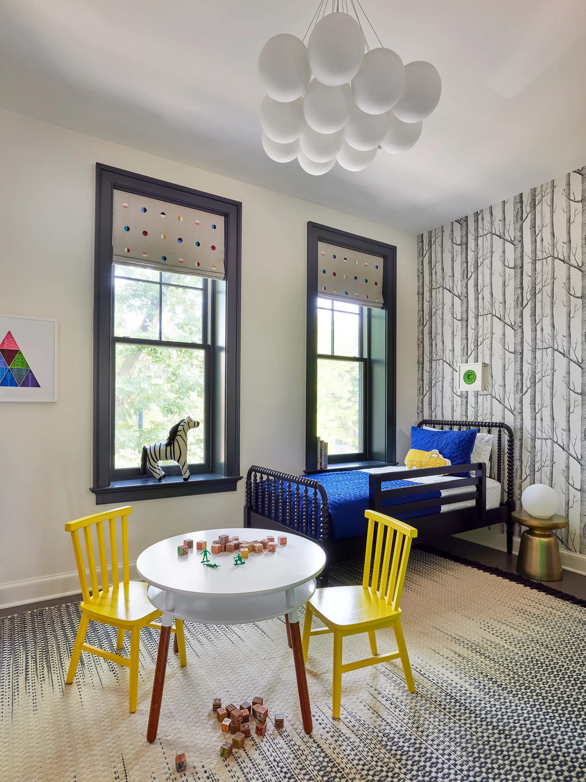 A children's bedroom features a black spool bed against tree-patterned wallpaper, a white play table with yellow chairs, and a cloud-like pendant light.