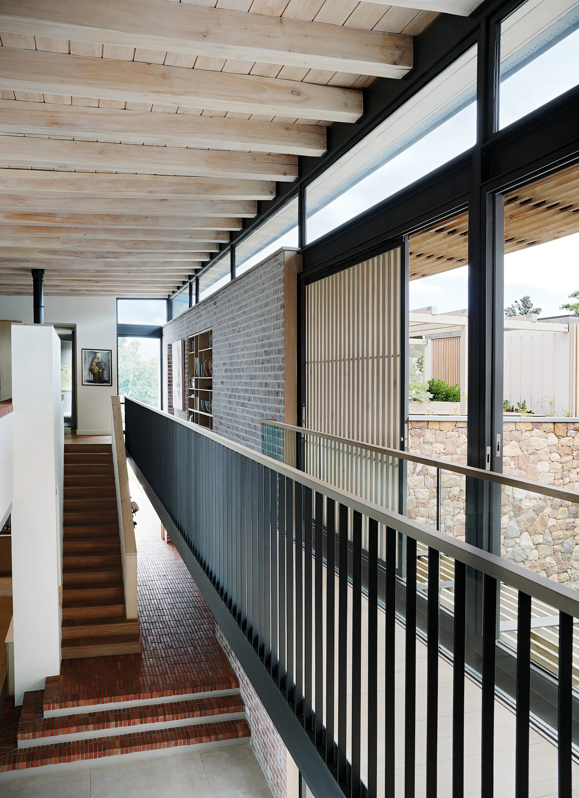 A modern hallway features a light wood beam ceiling, a long black metal railing, an exposed brick wall, and large windows looking outside.