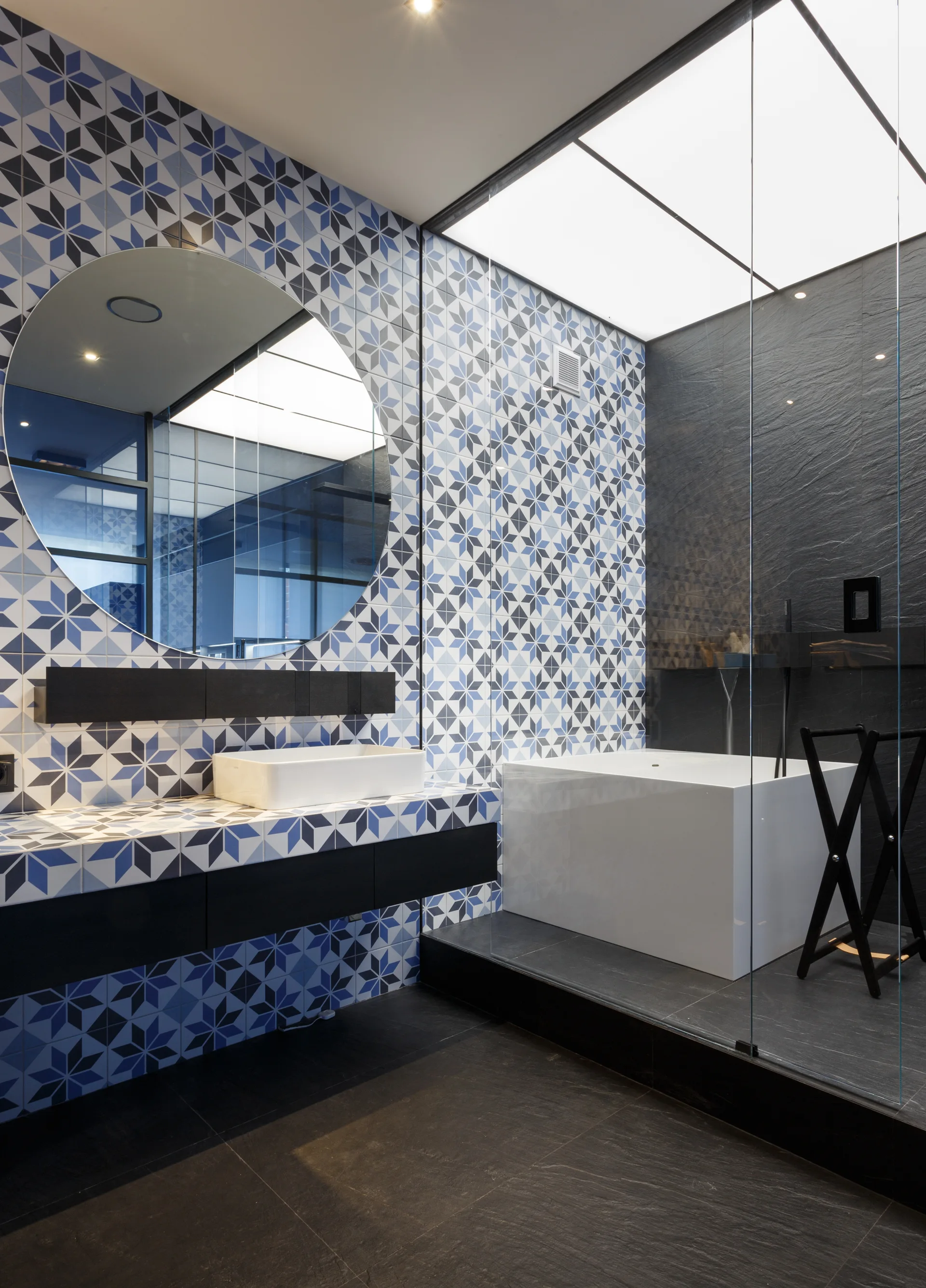A modern bathroom features blue and white geometric star-patterned wall tiles, a round mirror, a white vessel sink, and a glass-enclosed shower area.
