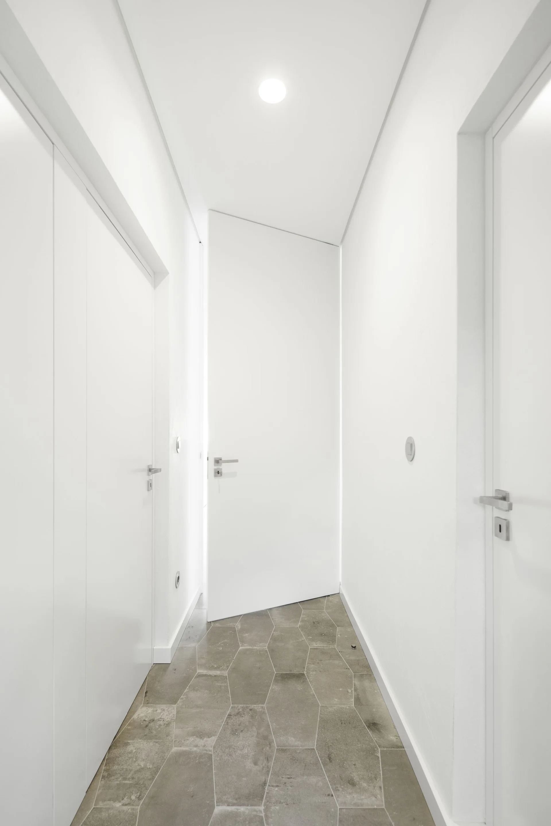 A narrow white minimalist hallway features sleek flush doors, silver handles, and a patterned floor of grey hexagonal stone-like tiles.
