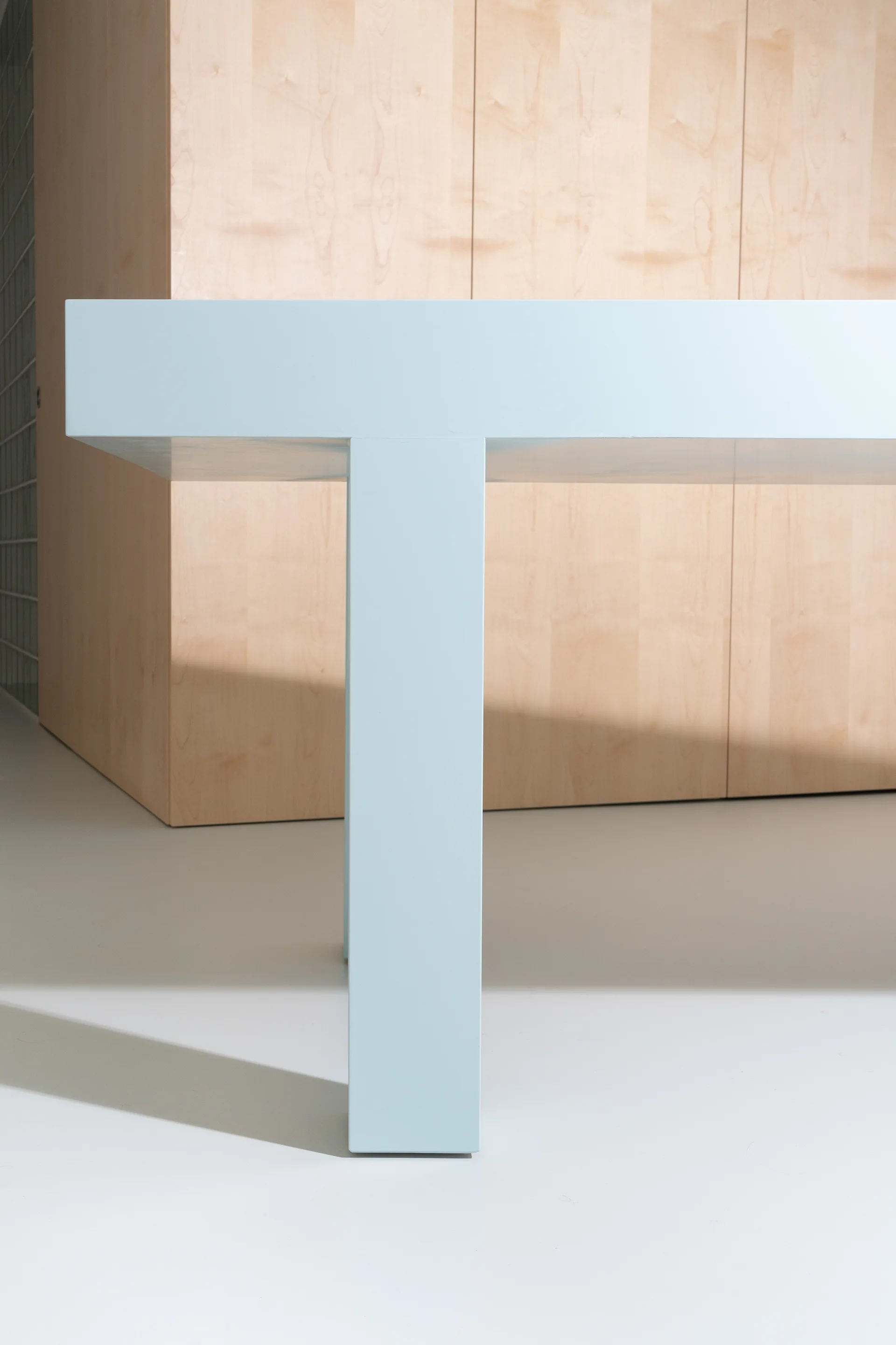 A close-up shot of a minimalist pale blue table leg and top positioned in front of natural light wood-paneled cabinets.