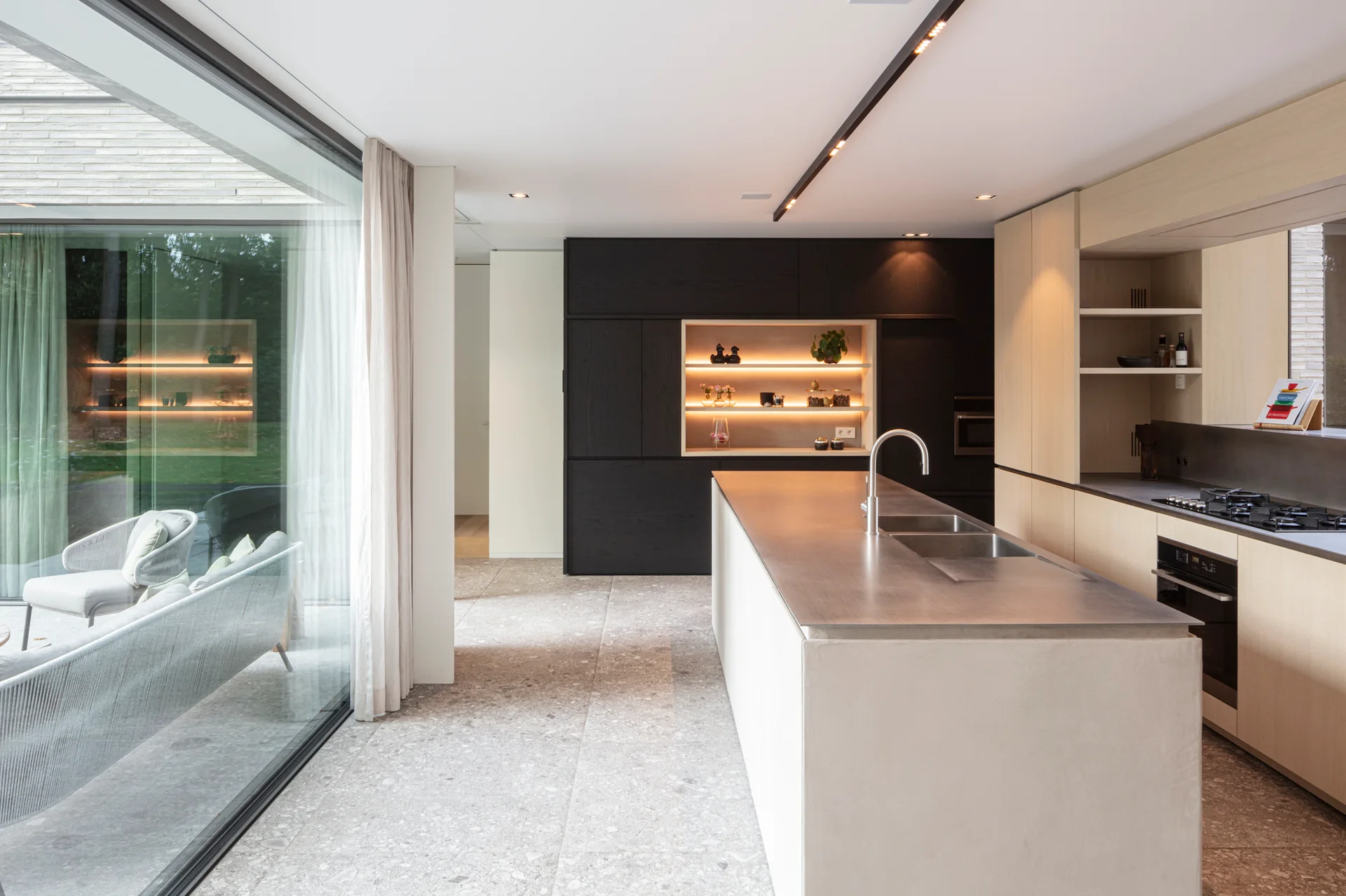 A minimalist kitchen features a central stone island, light wood and black cabinetry, and a sliding glass door opening to an outdoor patio.